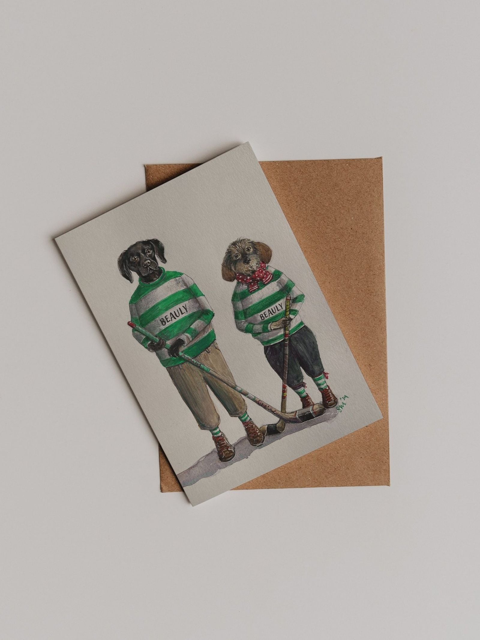 Send your warm wishes with the Beauly Shinty Players Greeting Card by Campbell’s of Beauly—a charming thank you note featuring Sophie Murray’s illustration of two dogs in green and white striped sweaters holding hockey sticks, beautifully presented on a classic brown envelope.