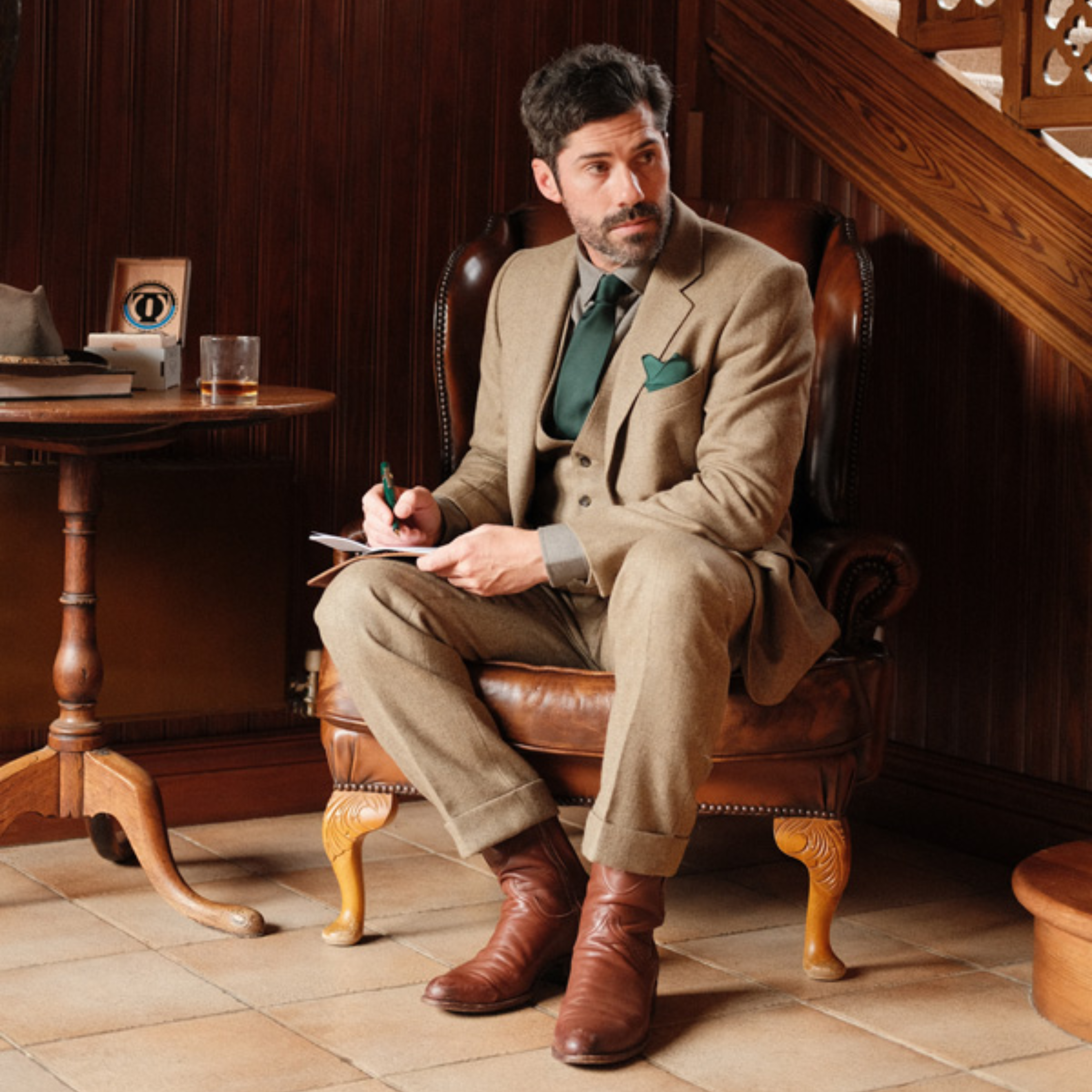 A man in a tan suit with a green tie and pocket square from Campbells of Beauly sits on a leather chair, writing in a notebook. He has dark hair, a beard, and brown boots. A drink, hat, and wooden furniture surround him in the warmly lit room.