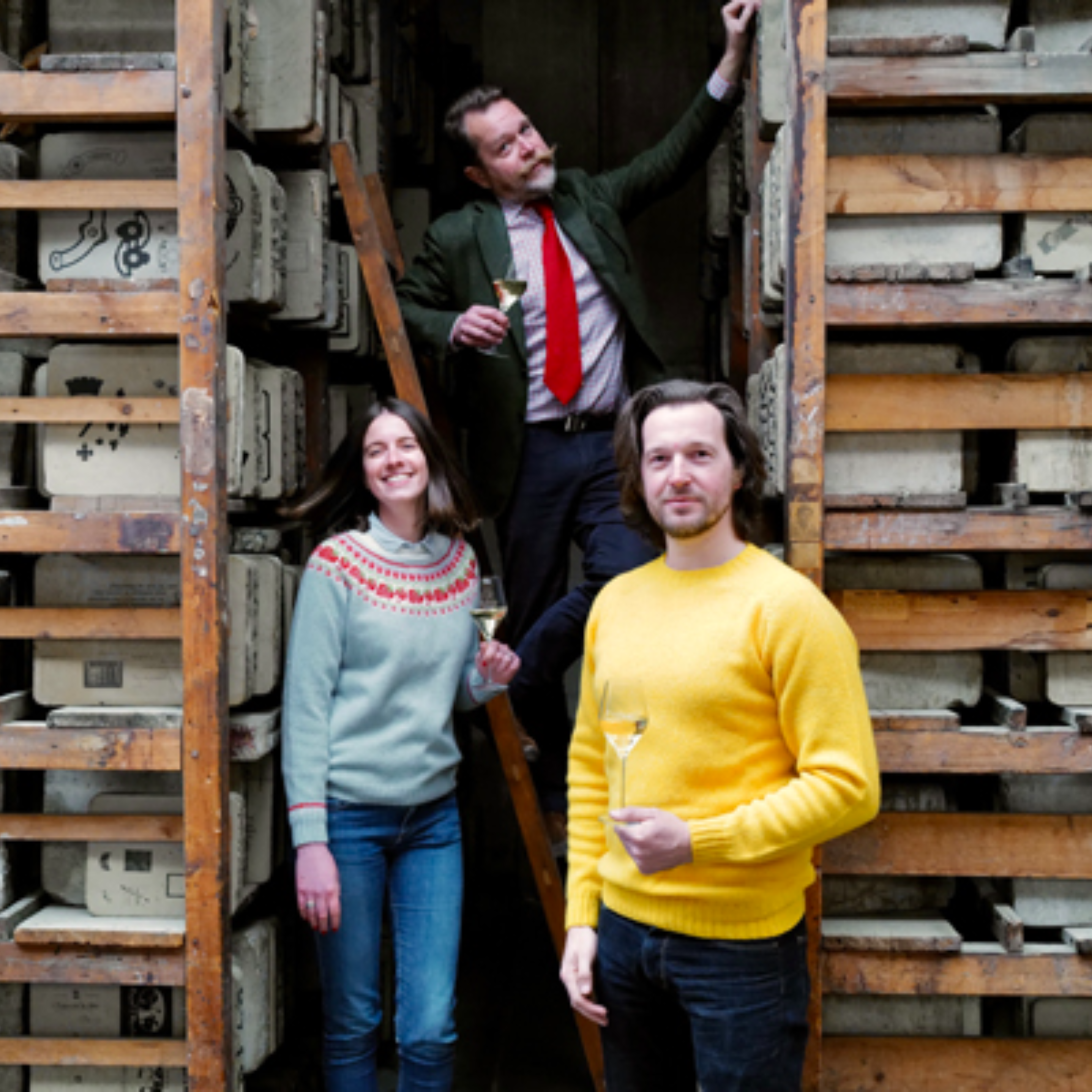 Three people holding glasses stand among shelves filled with stone slabs. One person is on a ladder wearing a green jacket, another in a grey sweater with red patterns, and the third in a yellow sweater stands in front.