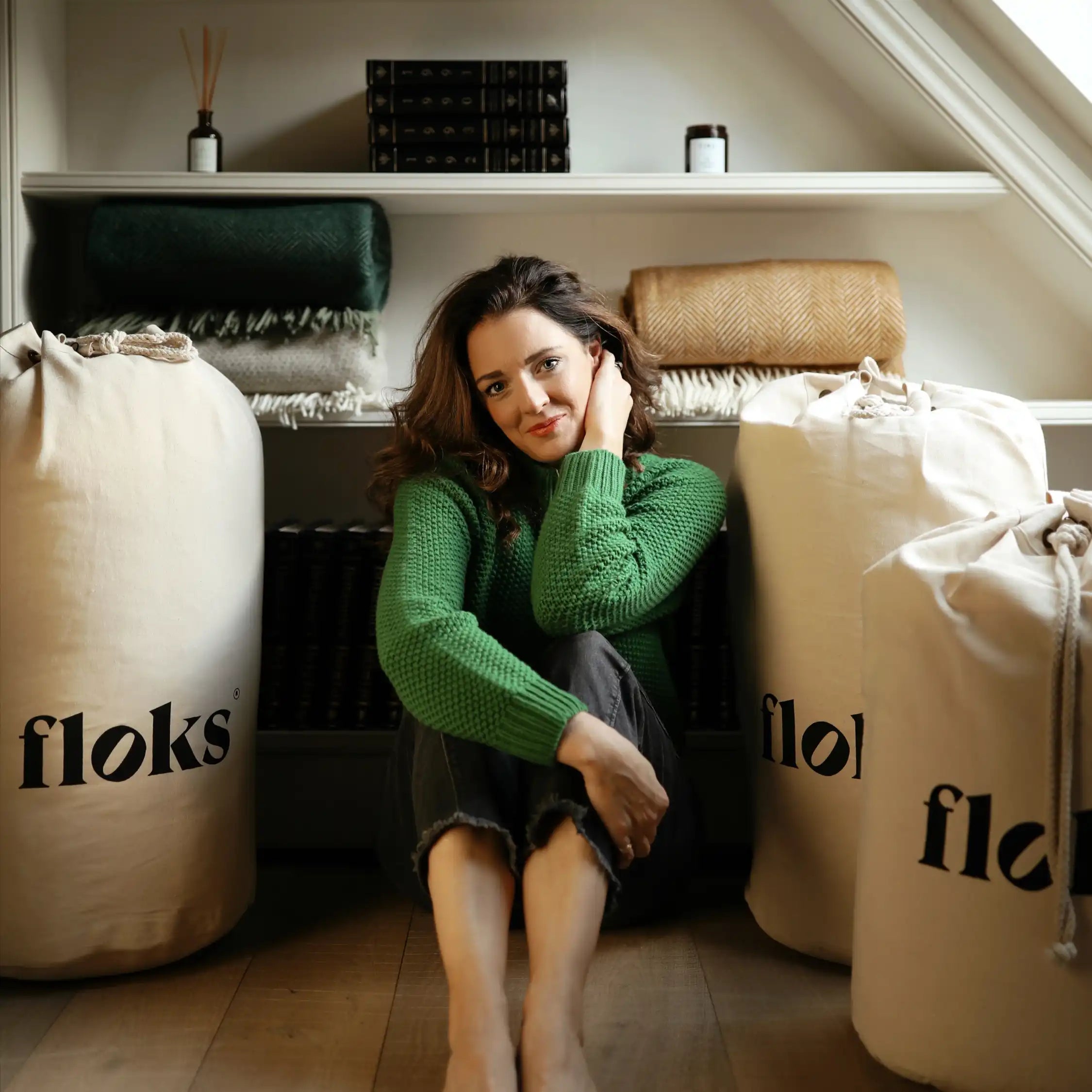 A woman in a green sweater from Campbells of Beauly sits barefoot on a wooden floor between two large canvas bags labeled floks, with shelves holding folded blankets and books behind her.