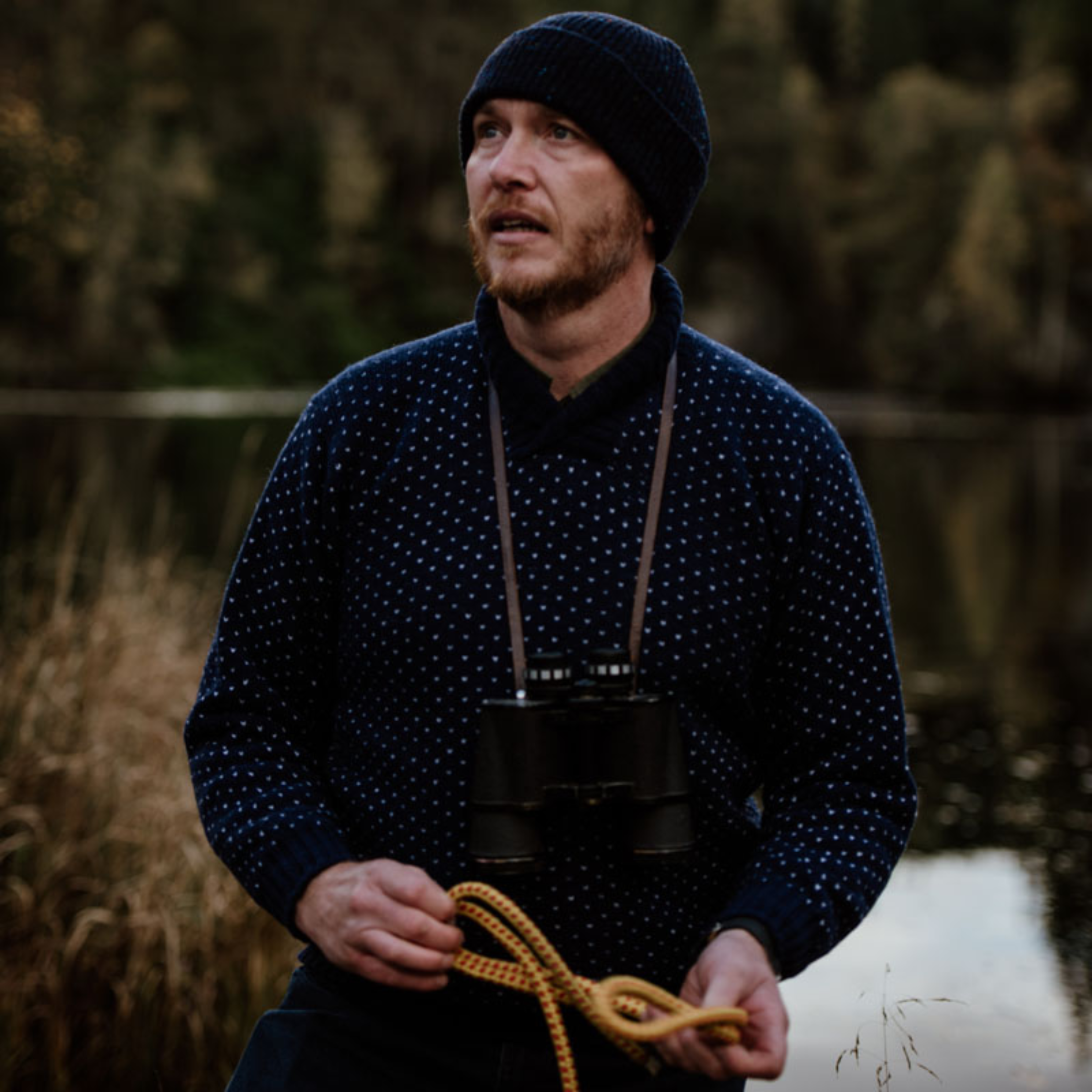 A man in a navy blue Campbells of Beauly sweater and beanie stands by a lake, holding a yellow rope with binoculars around his neck. Tall grass and trees are in the background.