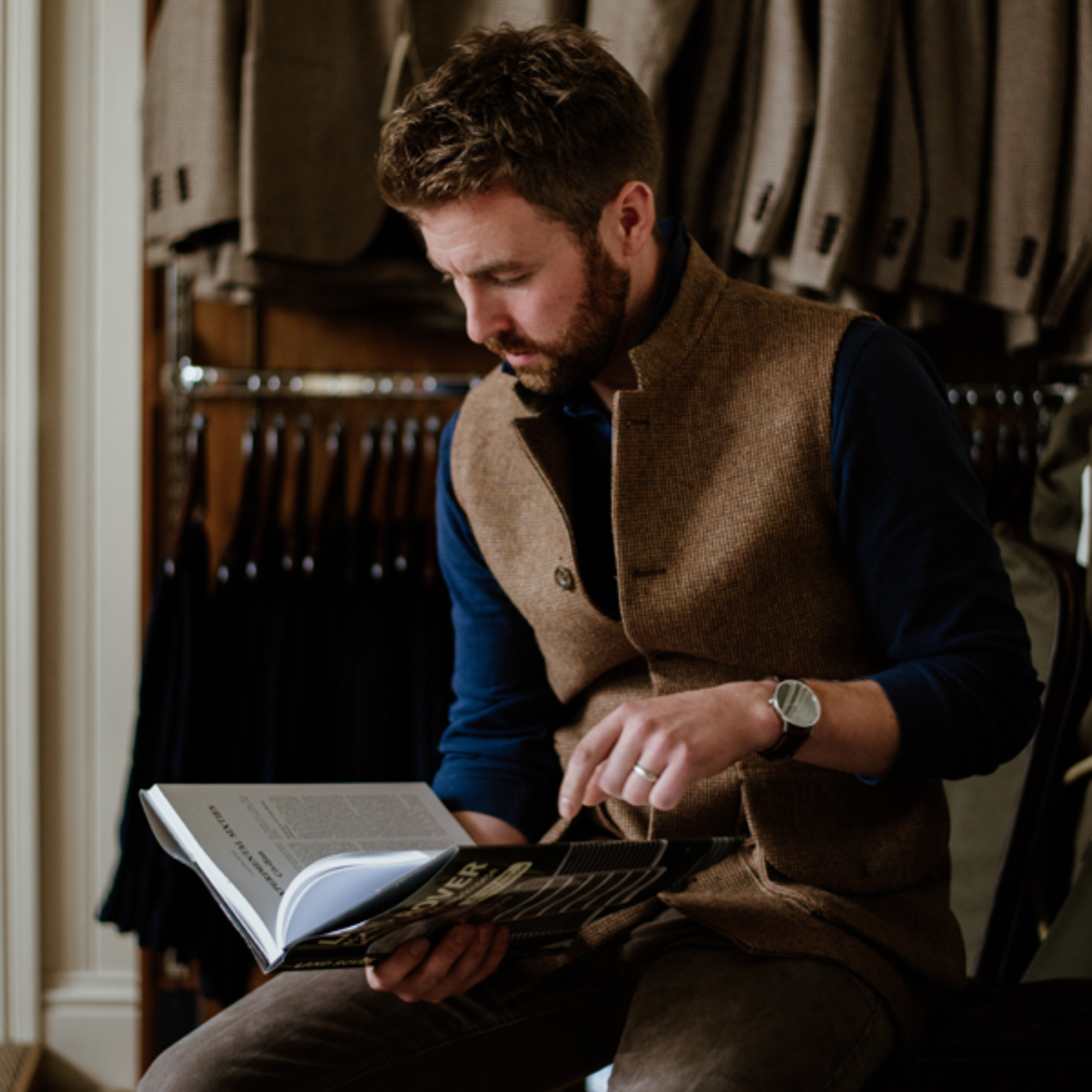 A man with a beard sits indoors, dressed in a brown wool vest and dark shirt, reading a book. Behind him, tailored jackets hang neatly on racks, suggesting a refined or boutique setting.