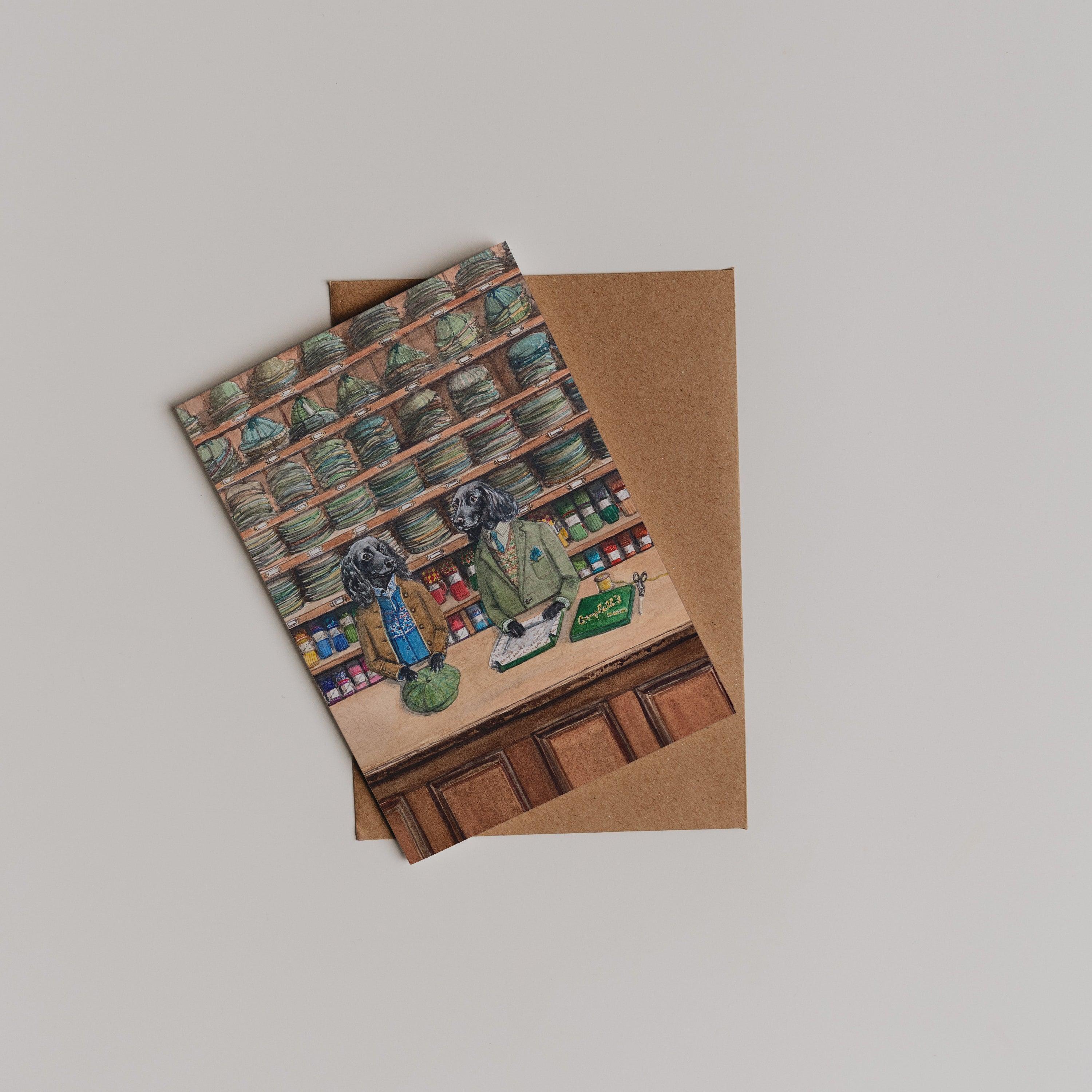 The Campbells Hats Greeting Card by Campbells of Beauly features two dogs in suits behind a shop counter, surrounded by jars and bottles. Perfect as a thank you note, it comes with a brown envelope on a light gray background.