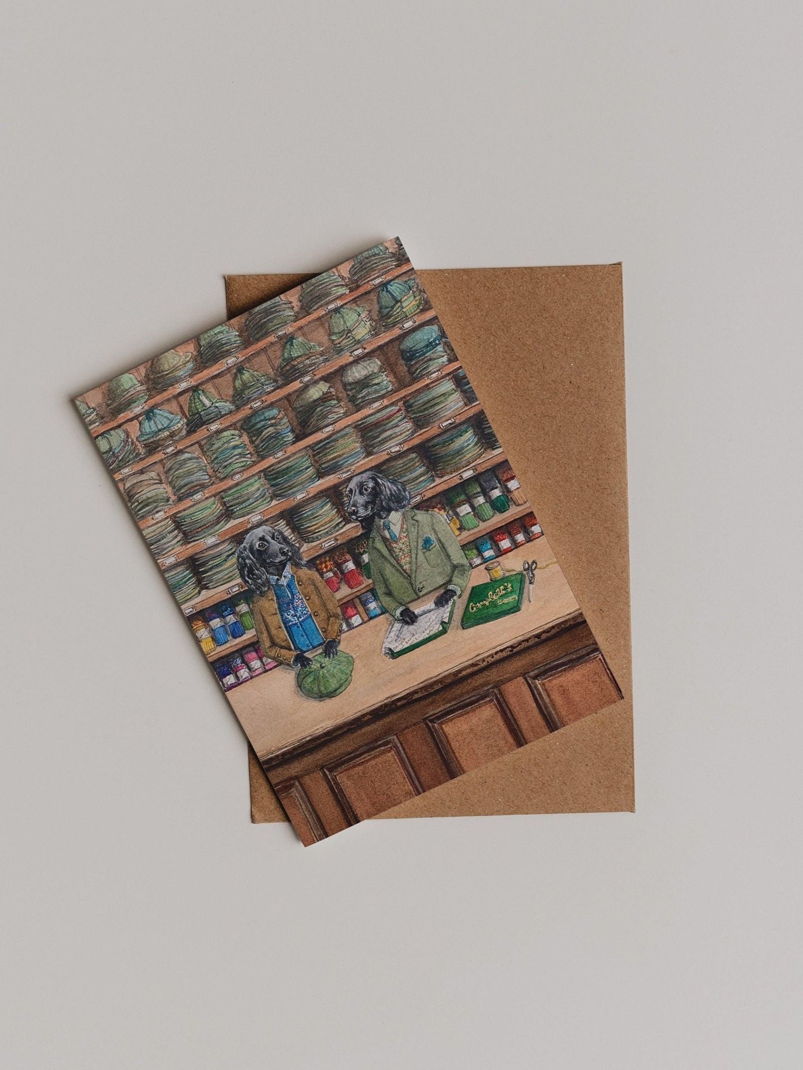 A delightful Campbell’s Hats Greeting Card from Campbell’s of Beauly, featuring two dogs dressed as shopkeepers behind a counter in a store lined with shelves of jars—perfect as a thank you note and presented on top of a brown envelope.