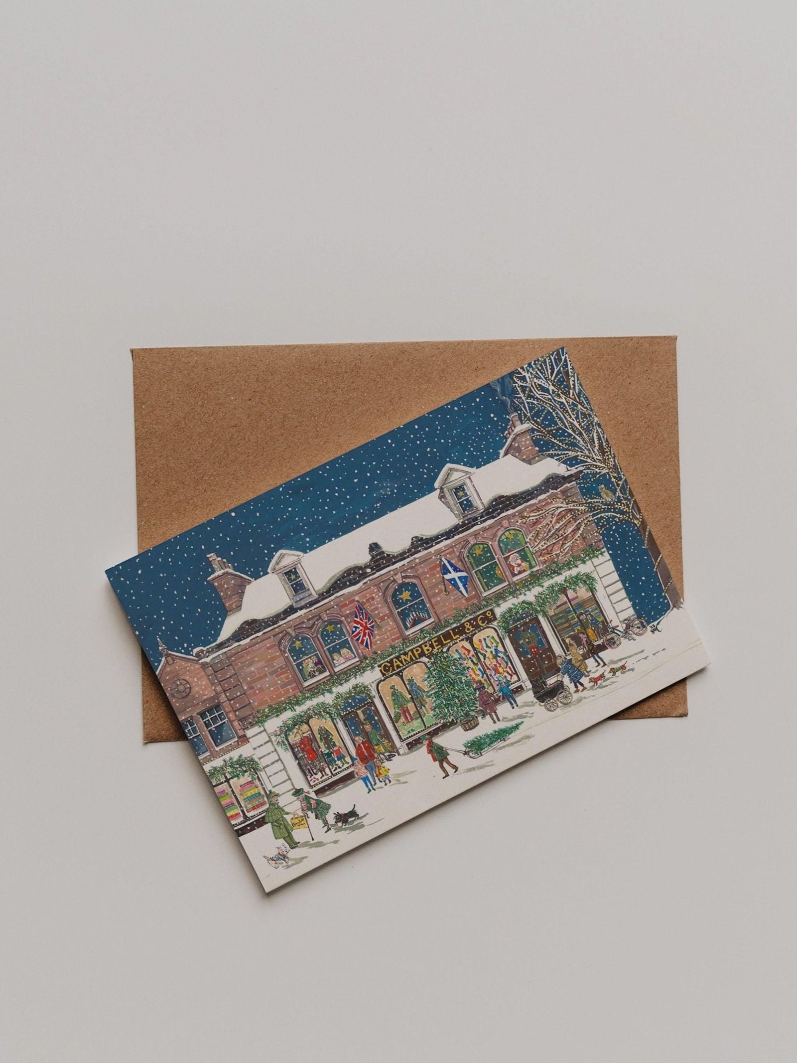 The Campbell’s at Christmas Greeting Card from Campbell’s of Beauly rests atop a brown envelope on a light surface, showcasing a snowy store scene and festive flags. The elegant illustration by Sophie Murray enhances the charm of this seasonal thank you card.