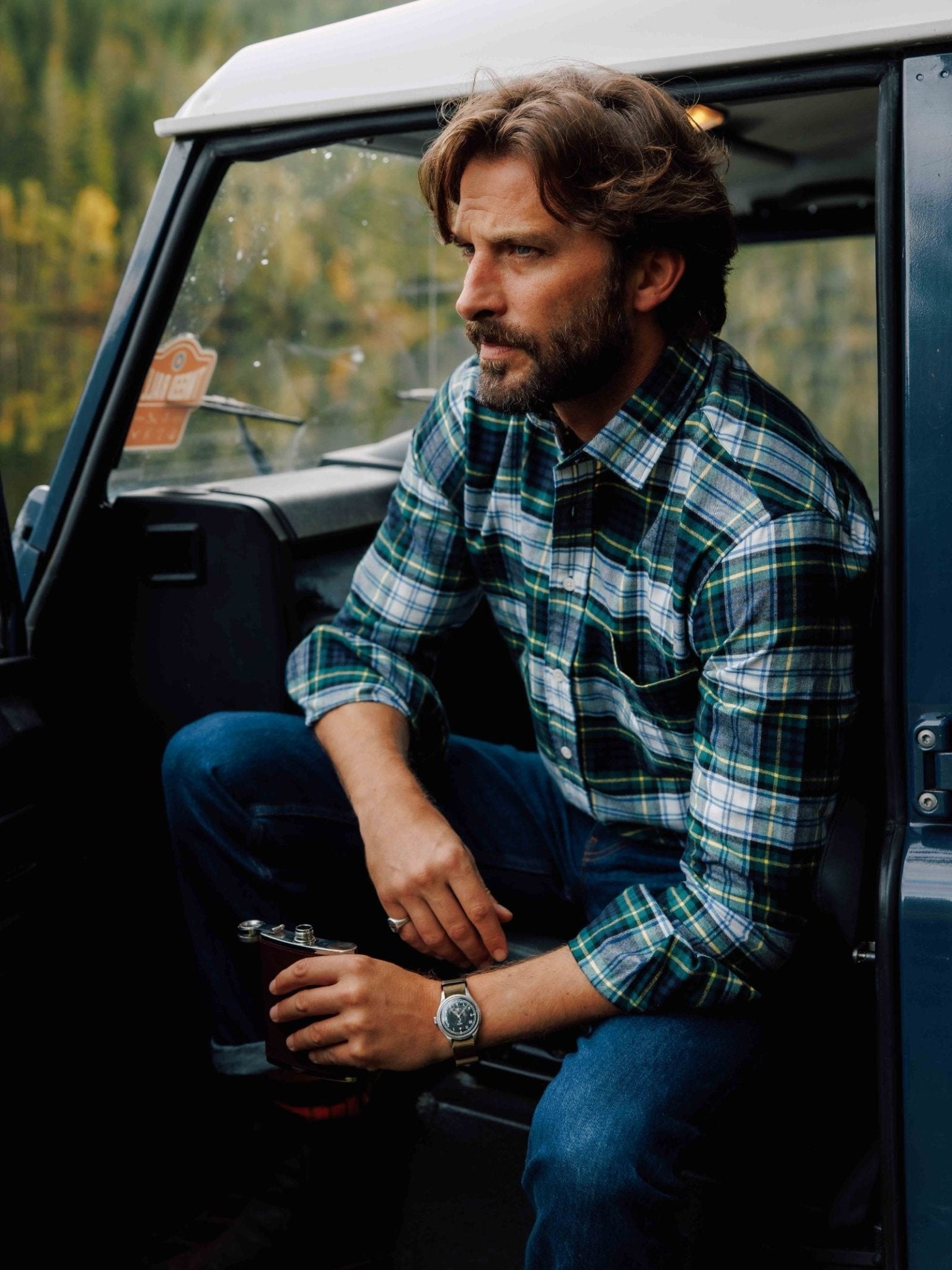 A man with wavy brown hair and a beard sits in a car doorway, wearing jeans and a Campbell's of Beauly Tartan Shirt, holding a metal flask and gazing thoughtfully into the distance.