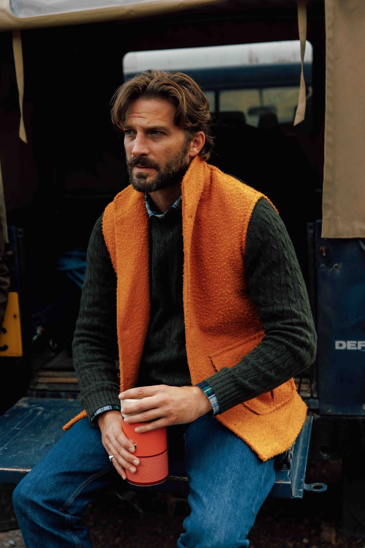 A man with wavy brown hair and a beard, dressed in a dark sweater, jeans, and a Campbell's of Beauly Casentino Fleece Gilet, sits on a vehicle’s back, holding a red travel mug and gazing thoughtfully to the side.