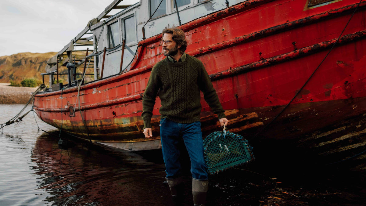 A man wearing a green cable-knit jumper and waders stands in shallow water, holding a wire lobster pot in front of a grounded red fishing boat.