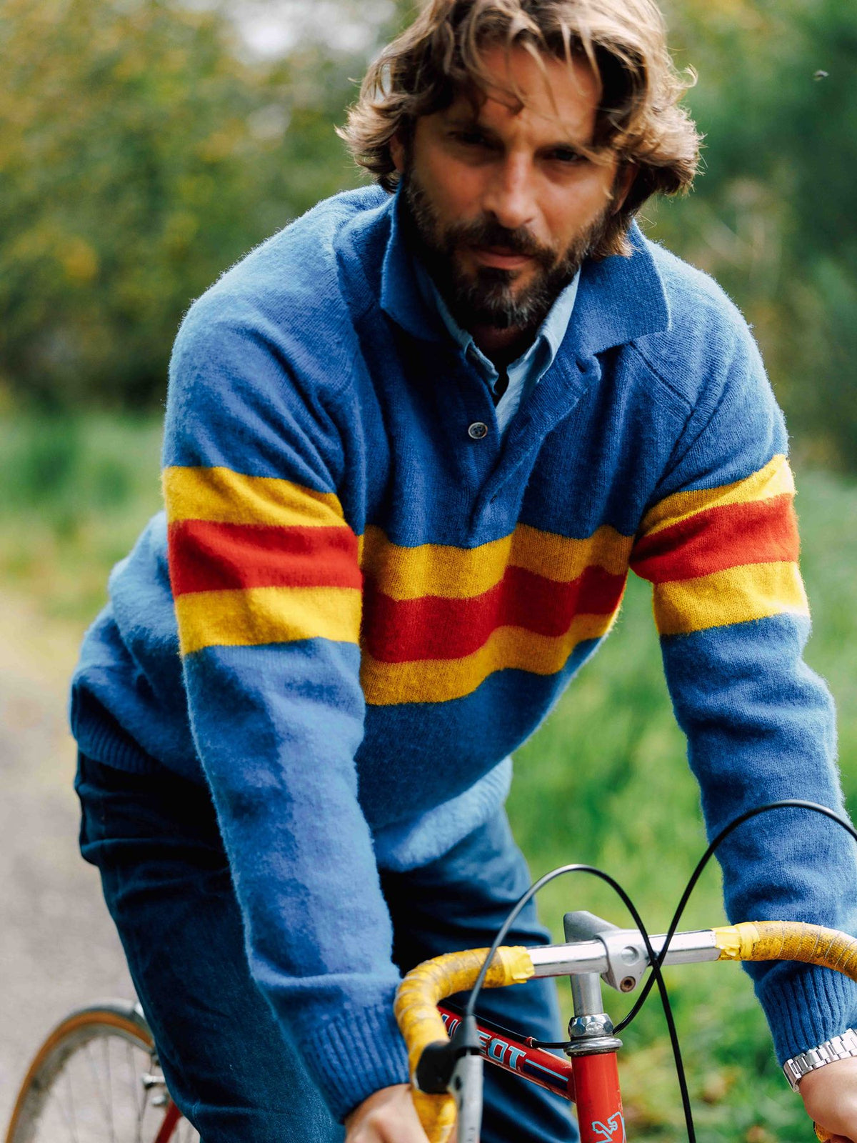 A bearded man wearing a blue jumper with horizontal yellow and red stripes rides a bicycle.