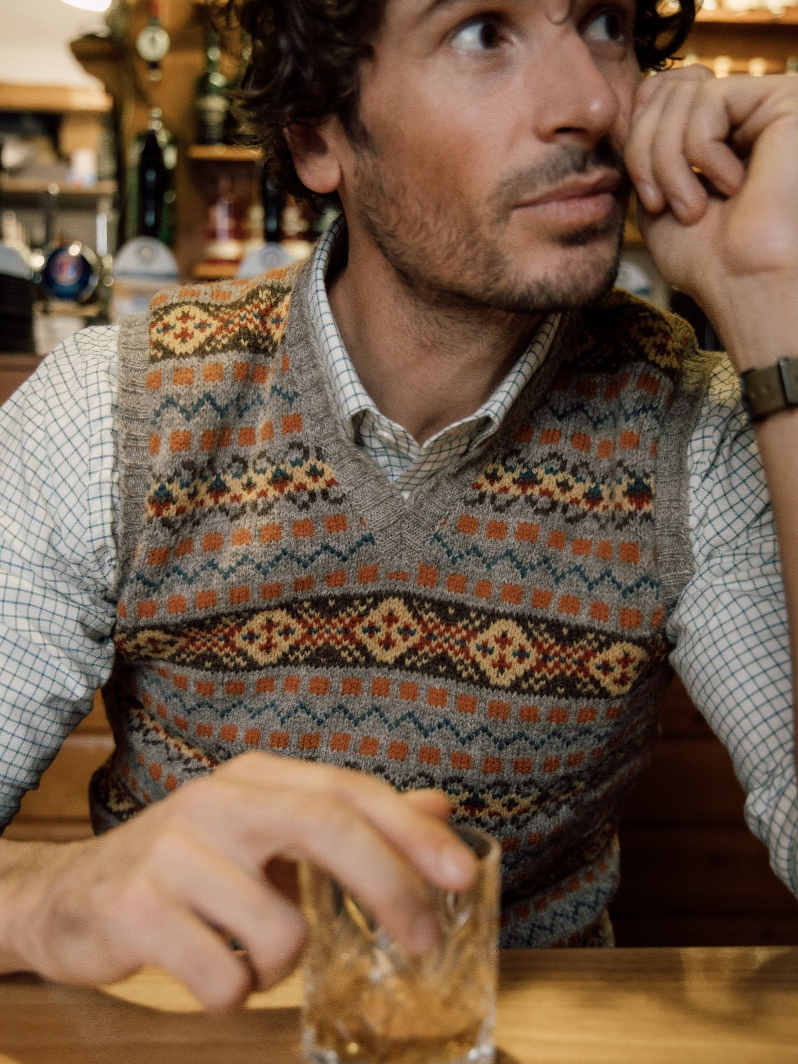 A man with curly brown hair and stubble wears a Campbells of Beauly Fairisle Slipover over a white grid-checked shirt, seated at a wooden table with a glass of amber liquid in a cozy wood-paneled bar.