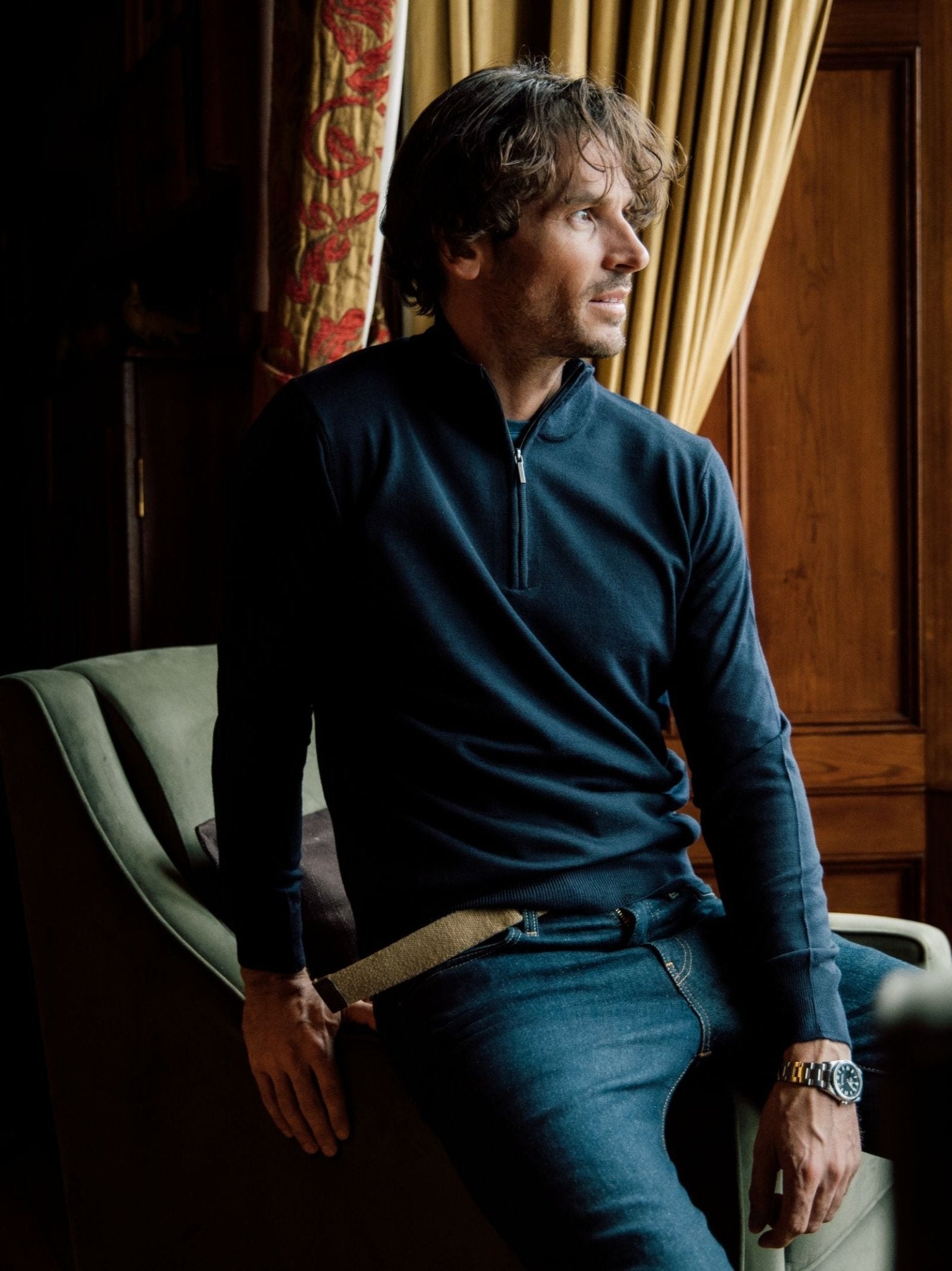 A man with wavy brown hair, wearing a dark Merino & Cashmere Quarter Zip Jumper from Campbells of Beauly and jeans, sits on the arm of a green chair by a window with gold curtains, looking thoughtfully to the side.