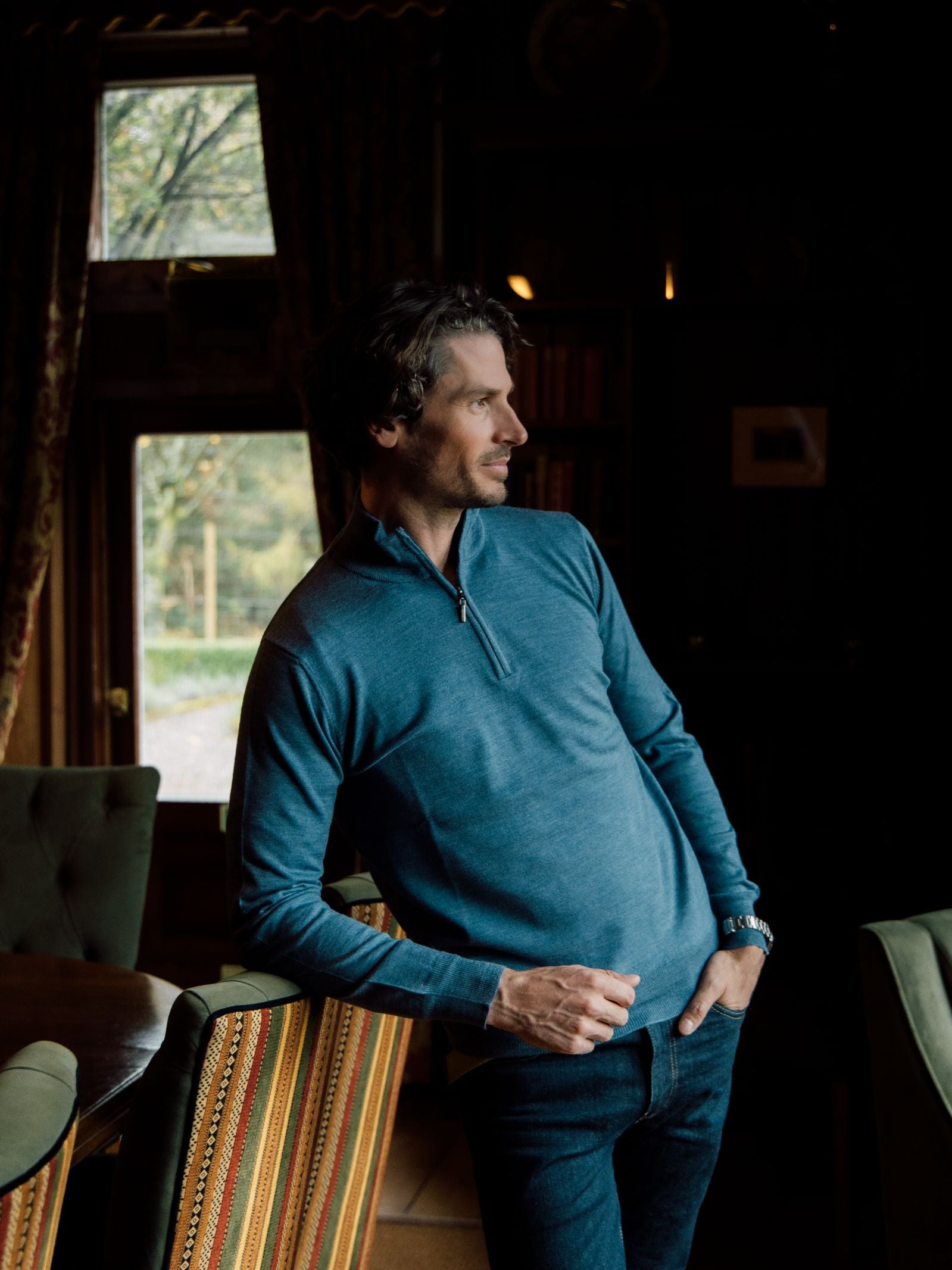 A man with wavy dark hair and light skin, dressed in a Campbells of Beauly Merino Quarter Zip Jumper and jeans, leans against a table in a dim room with patterned chairs, wood paneling, and large windows overlooking greenery.