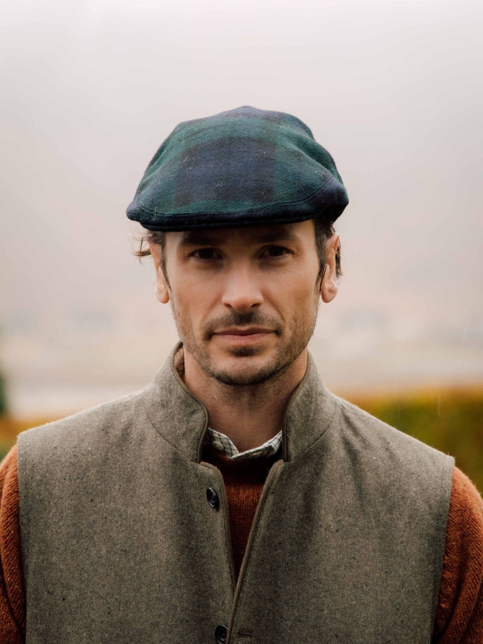A man with light skin and short brown hair wears a Campbell's of Beauly Blackwatch Tartan Conon Cap, a brown sweater, and a gray vest, standing outdoors against a foggy, blurred background.