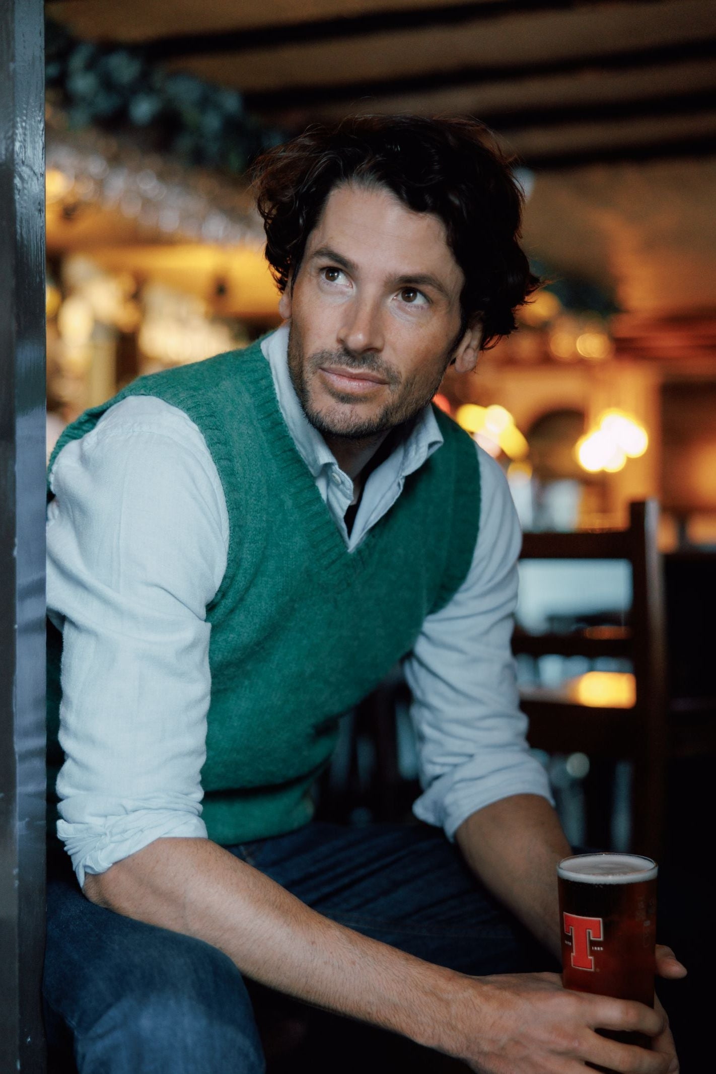 A man with wavy brown hair wears a Campbells of Beauly Shetland Slipover and sits in a dimly-lit pub, holding a pint of beer while looking thoughtfully to the side.