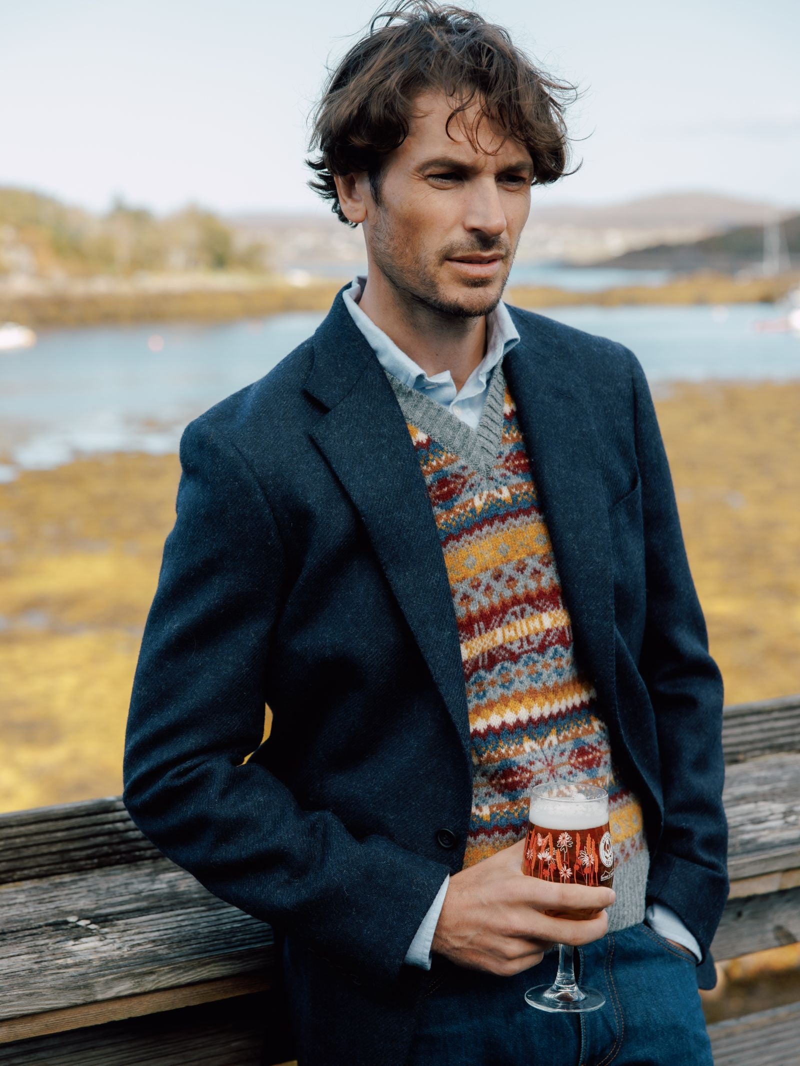 A man with wavy brown hair stands outdoors by water and trees, wearing Campbells of Beauly Navy Twill House Jacket over a patterned sweater vest and blue shirt. Holding a glass of amber beer, he gazes thoughtfully to the side.