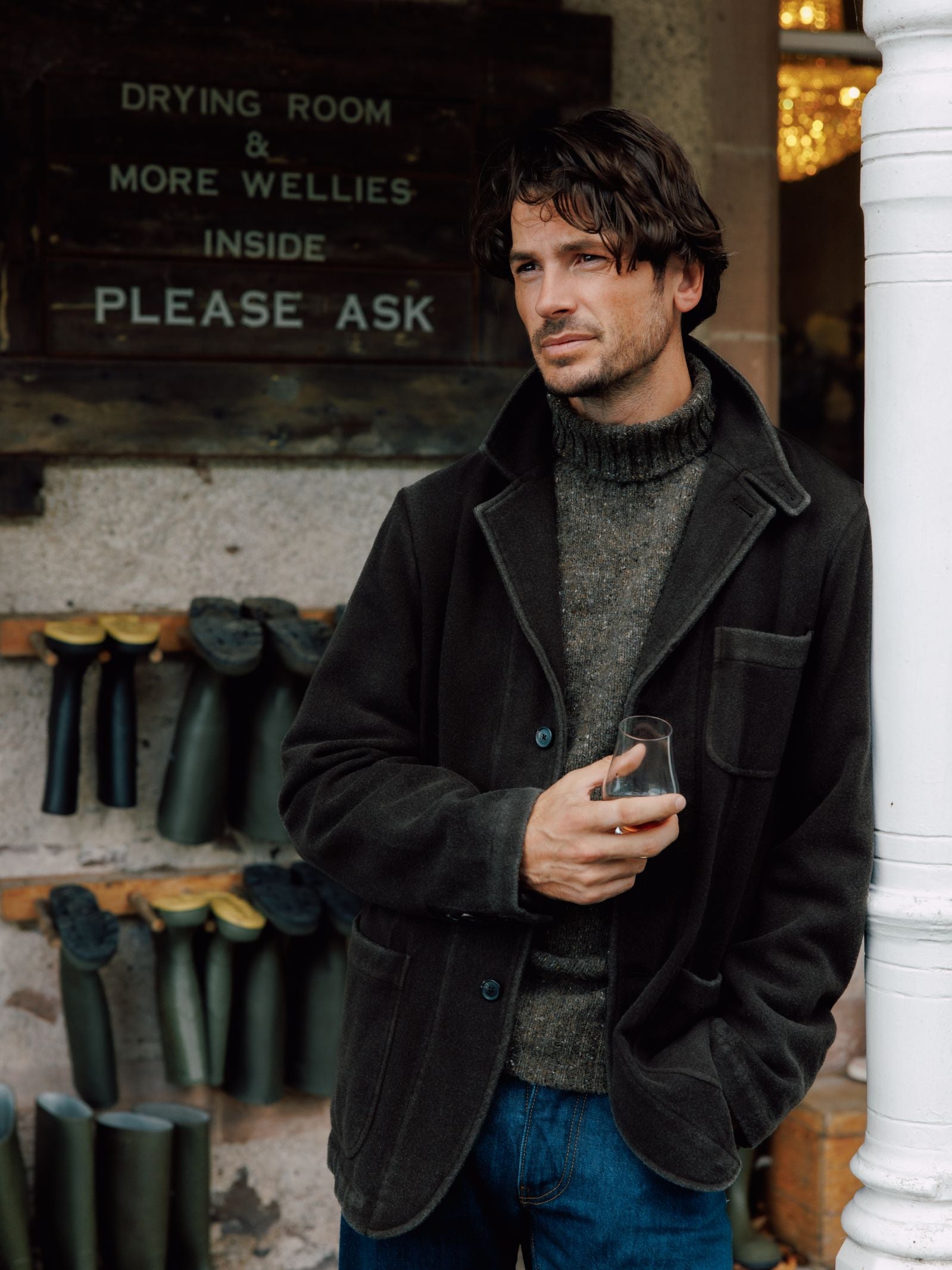A man with wavy brown hair and stubble stands outside, holding a glass. He wears jeans and a Campbells of Beauly Merino & Cashmere Jacket. Behind him is a rack of rubber boots and a sign: “Drying Room & More Wellies Inside Please Ask.”.
