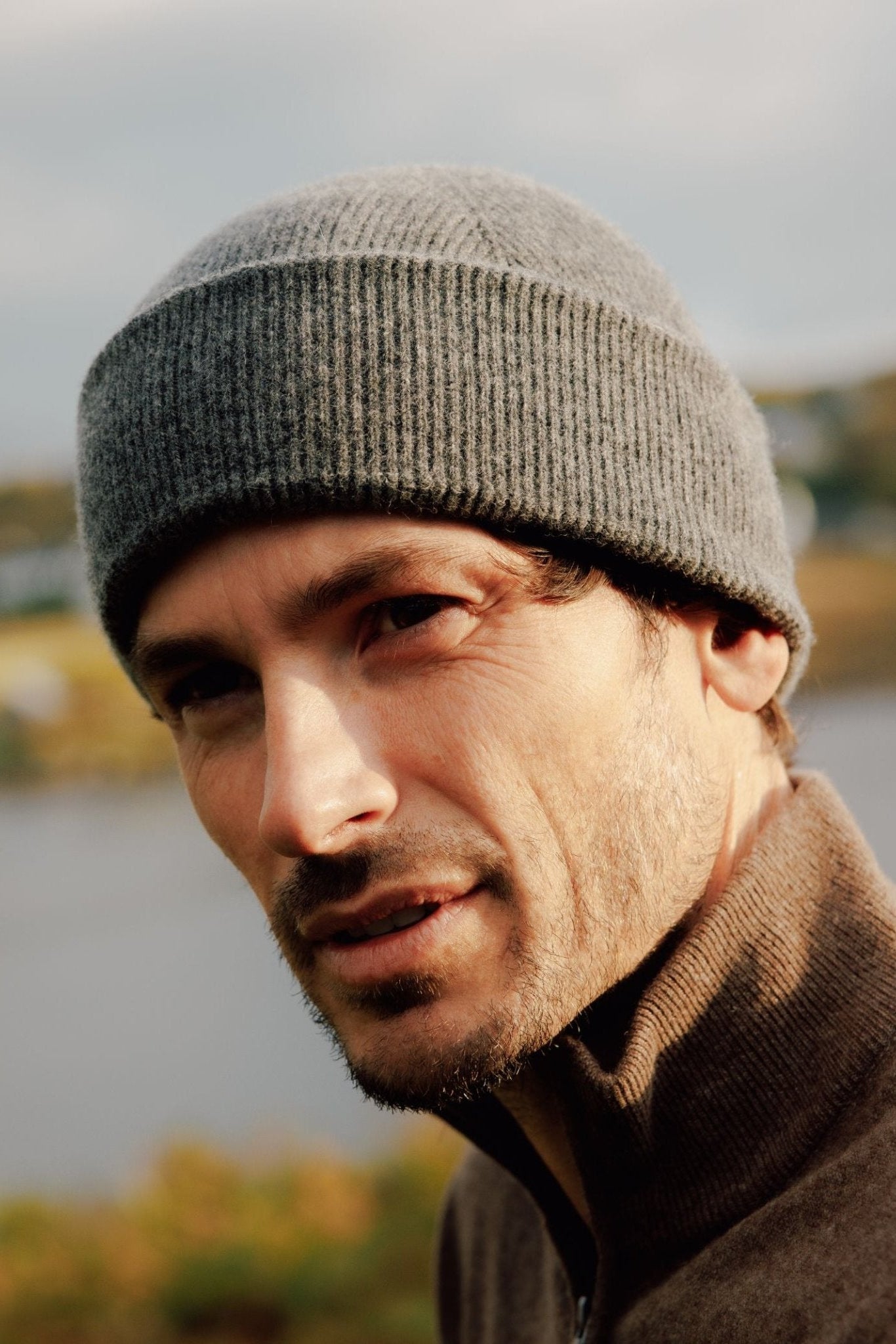 A man with light facial hair wears a gray Watchman Hat by Campbells of Beauly and a brown zip-up sweater, looking at the camera outdoors in the Scottish Borders with water and greenery blurred in the background.