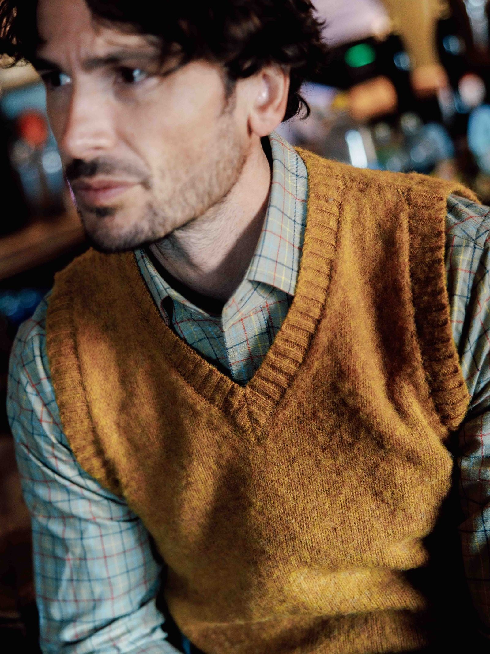 A man with wavy brown hair and light stubble, gazing left, wears a yellow knit vest over a Campbells of Beauly Windowpane Shirt. The softly blurred background suggests a warm, cozy indoor atmosphere.
