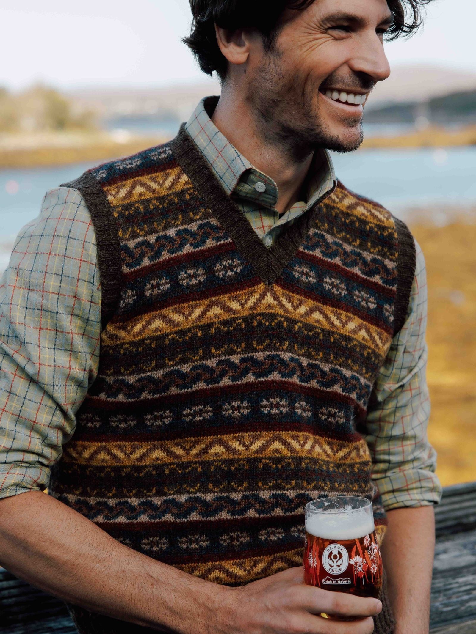A smiling man with wavy dark hair wears Campbells of Beaulys Fairisle Slipover over a checked shirt, standing by water with a pint of amber beer in hand and a blurred landscape in the background.