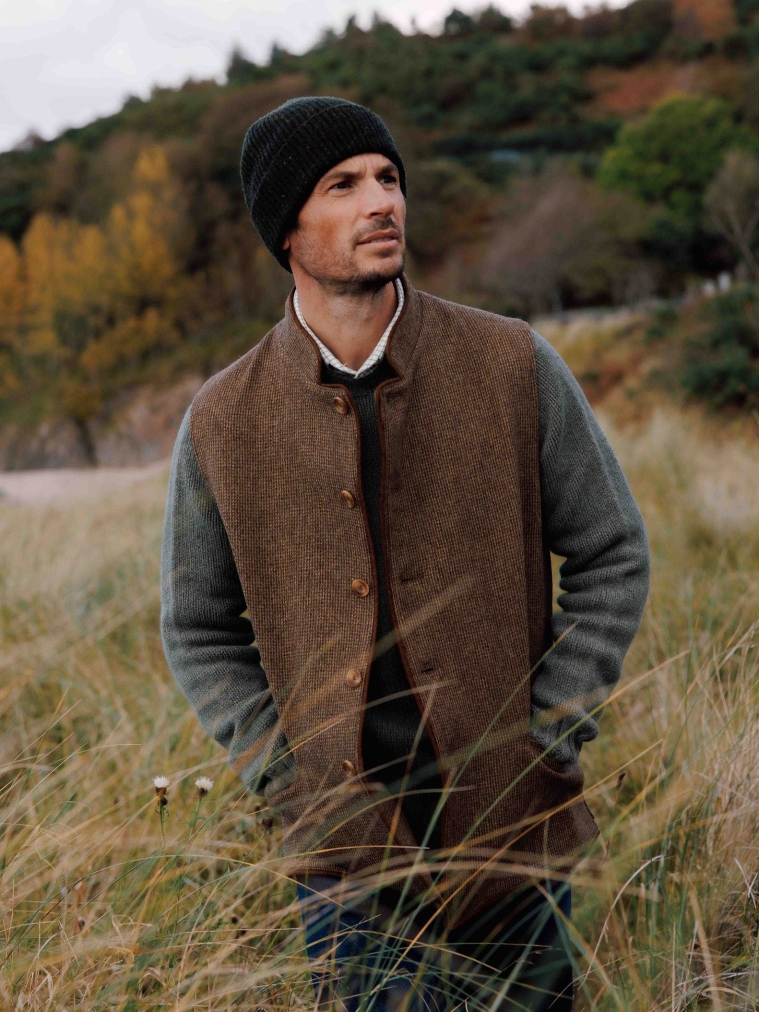 A man in a Campbell's of Beauly Tweed Nehru Gilet, green sweater, and black beanie stands in tall grass with trees and hills behind him, gazing thoughtfully into the distance.