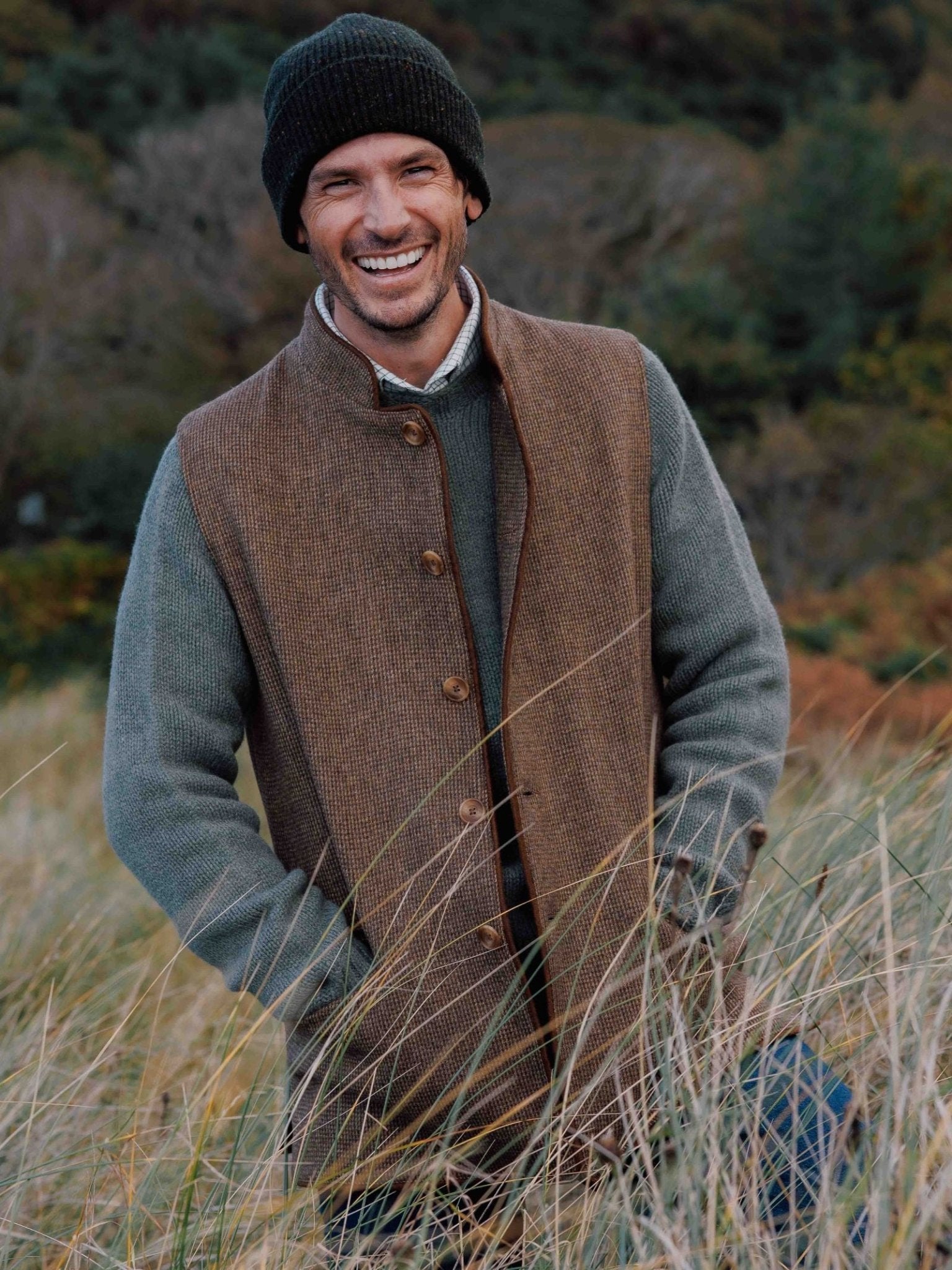 A smiling man wears a black beanie, gray sweater, and Campbell's of Beauly Tweed Nehru Gilet as he stands outdoors in tall grass with trees behind him.