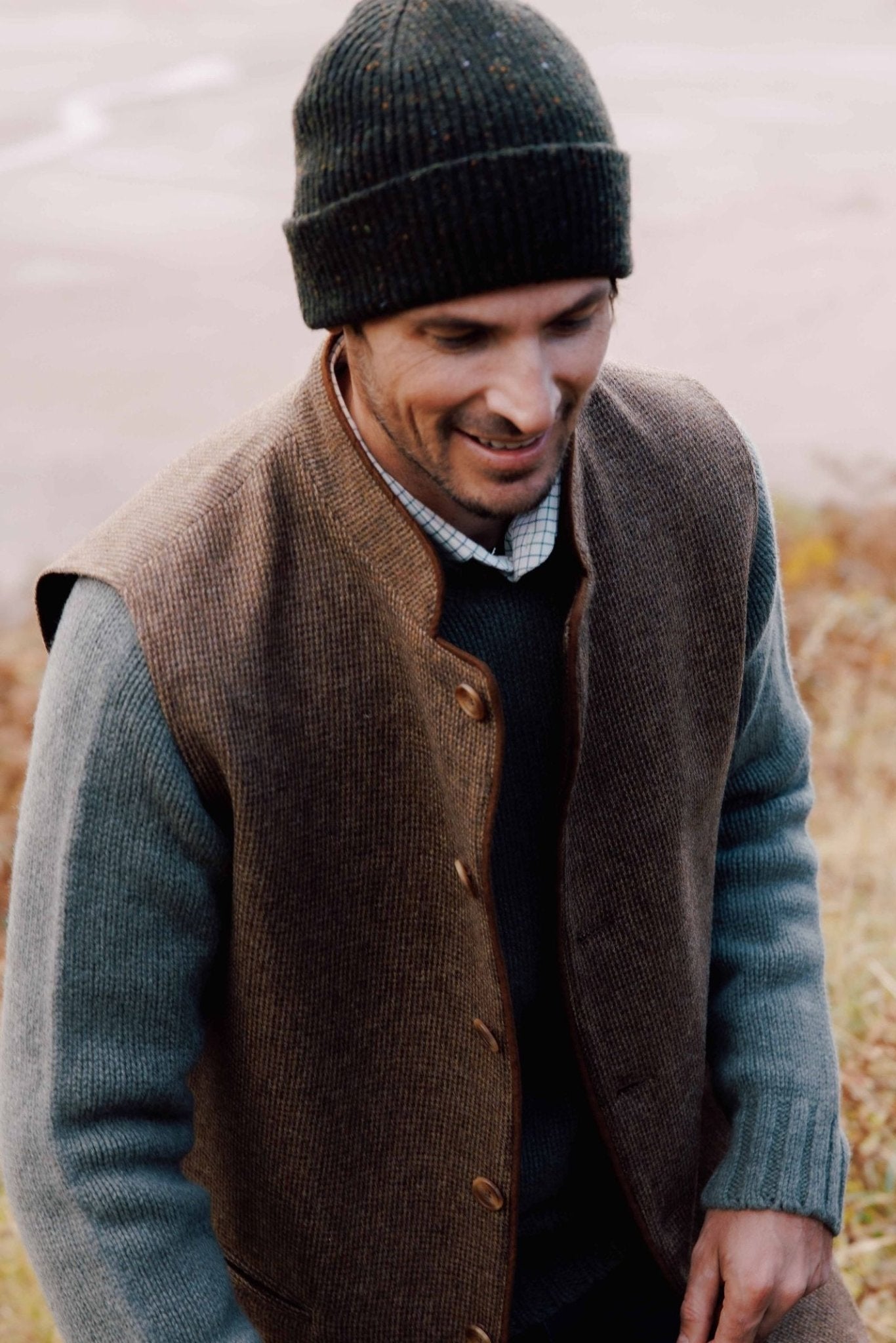 A man smiles outdoors on grass, wearing a Campbell's of Beauly Tweed Nehru Gilet, a gray sweater, and a dark knit beanie, with a blurred background.