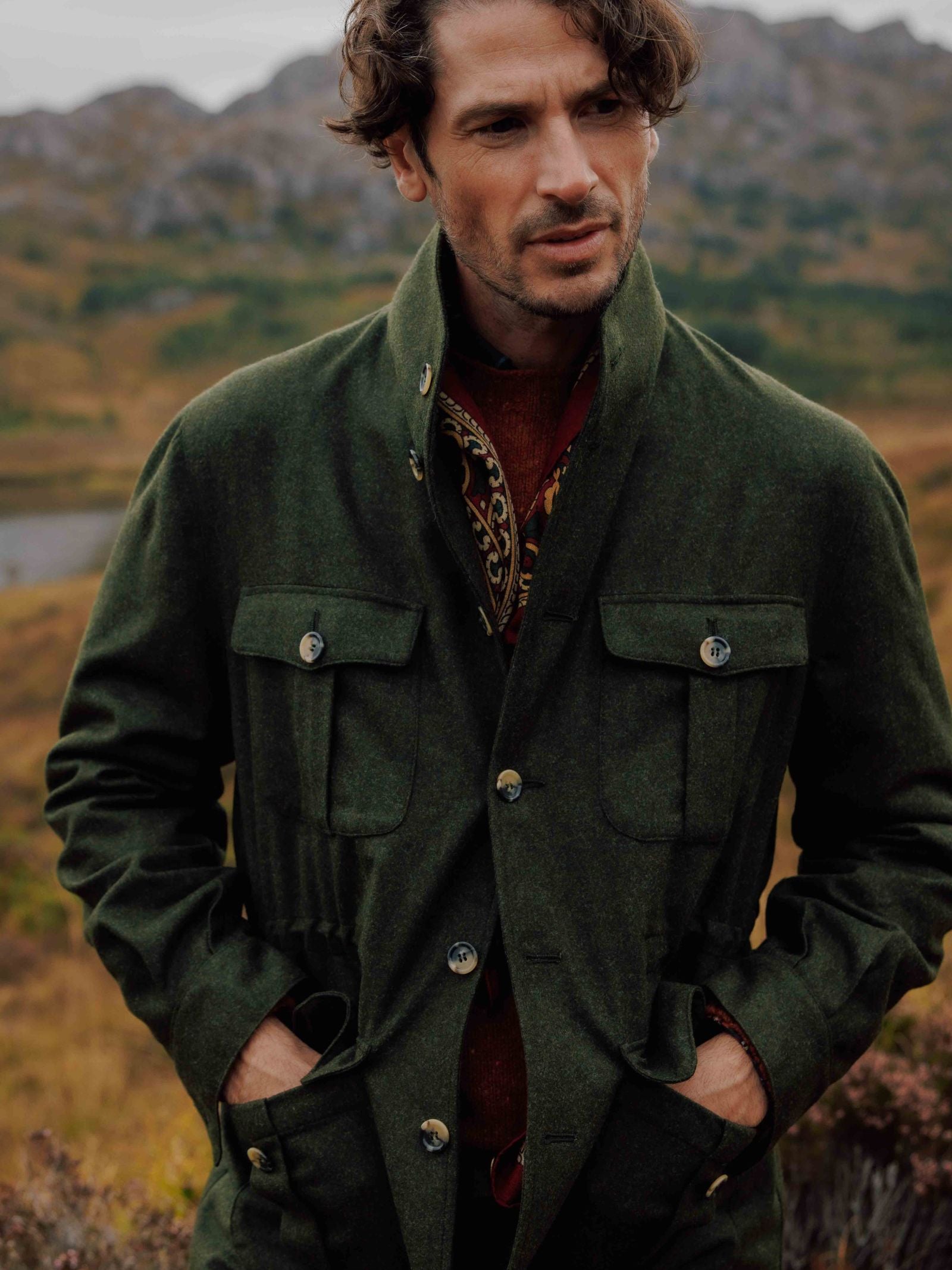 A man with wavy brown hair and a slight beard stands outdoors in green and brown hills under a cloudy sky, wearing the Campbells of Beauly Field Teba Jacket featuring flap pockets, with his hands in his pockets.