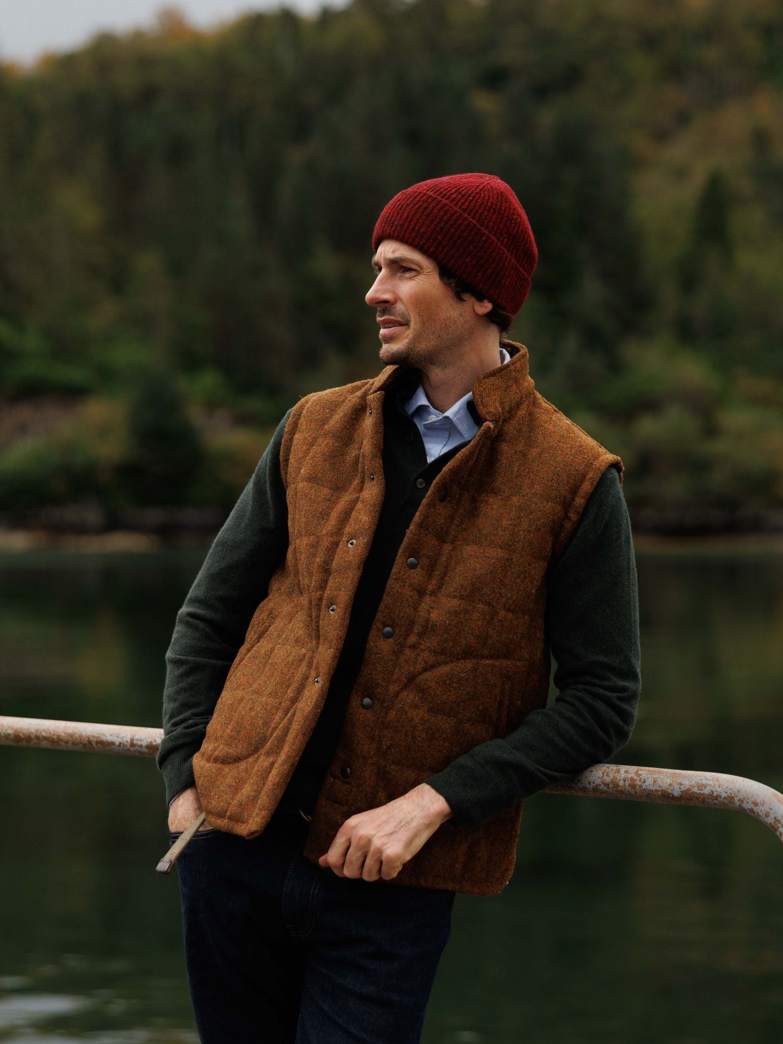 A man in a maroon beanie, dark green sweater, blue shirt, and the Harris Tweed Quilted Gilet by Campbell's of Beauly leans on a railing outdoors with a lake and forested hills behind him.