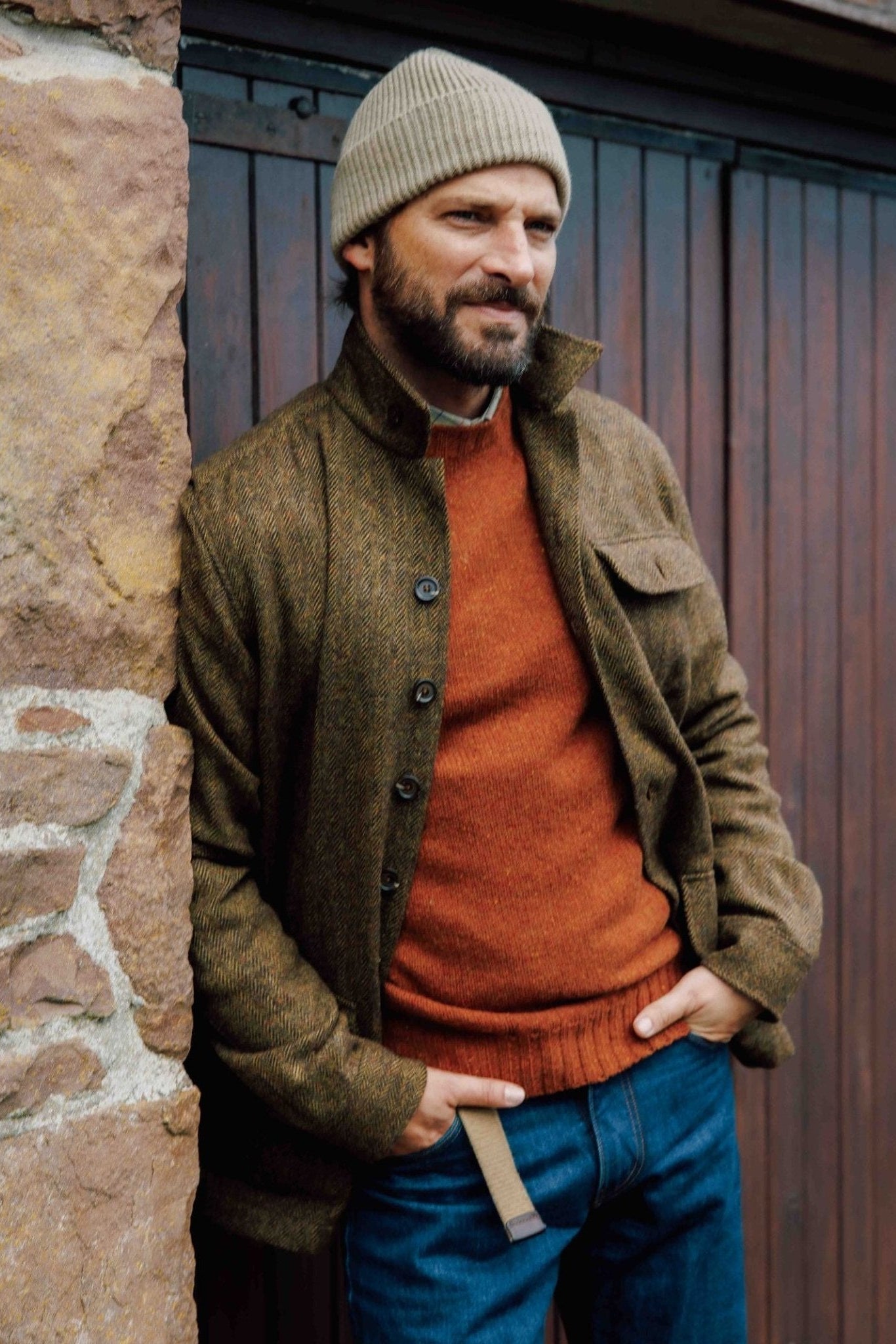 A bearded man in a beige knit beanie, brown jacket, blue jeans, and a Campbell's of Beauly Donegal Lambswool Crew Neck Jumper stands by a stone wall with hands in pockets, looking to the side.