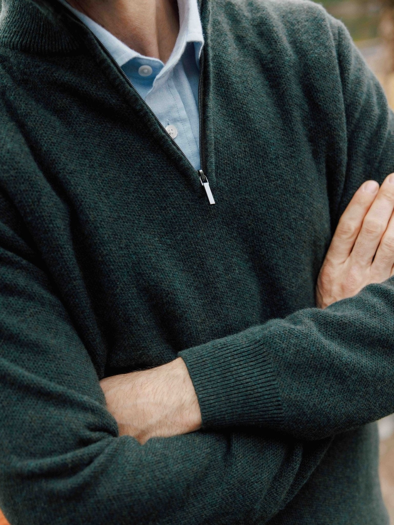 A person, face not visible, stands with arms crossed wearing a Campbell's of Beauly Piquet Quarter Zip Jumper in dark green over a light blue collared shirt.