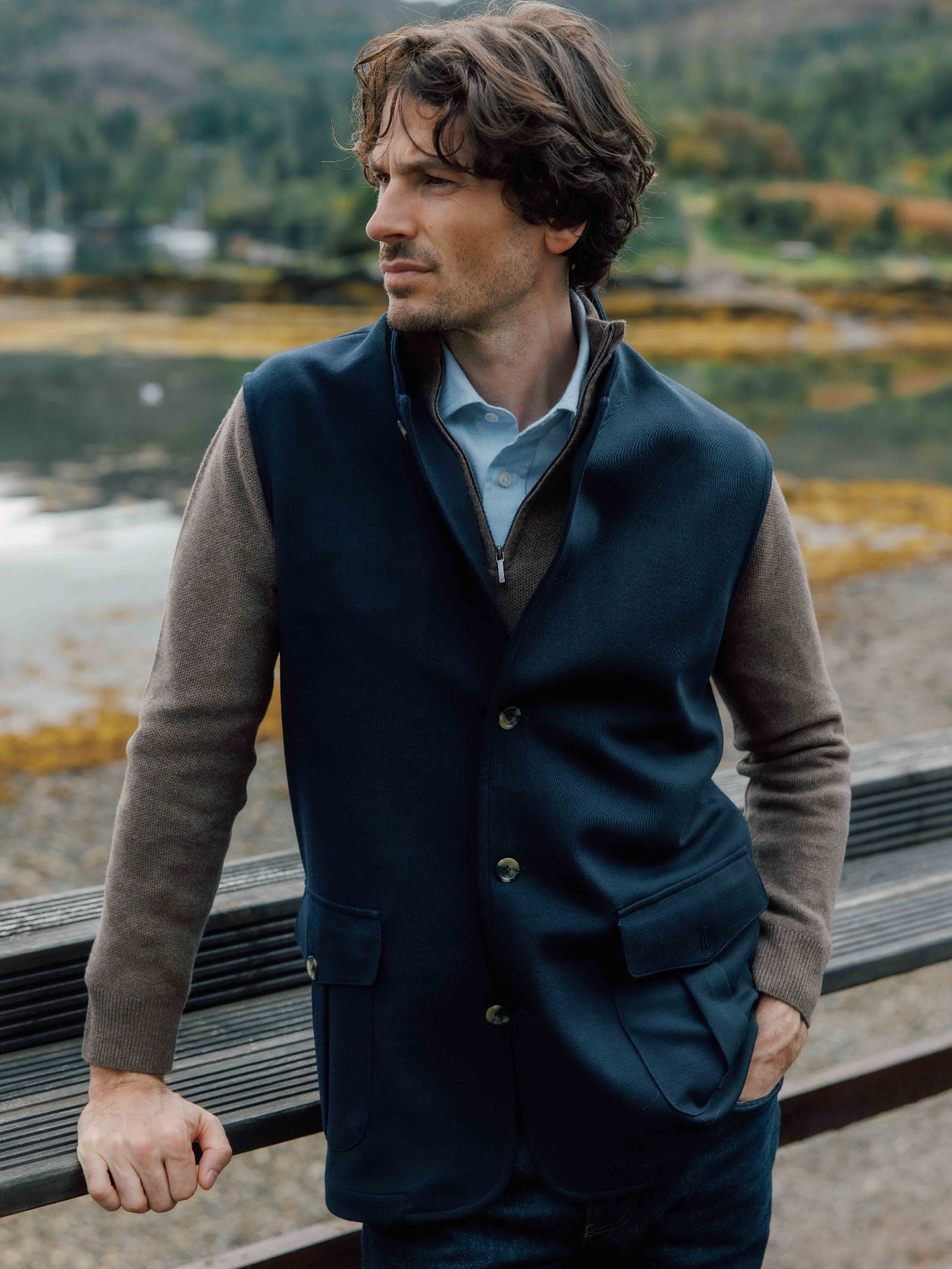 A man with wavy brown hair stands outdoors by water, wearing the Campbell's of Beauly Knitted Nehru Gilet over a brown sweater and light blue collared shirt, looking to the side with one hand in his pocket; trees and hills are behind.