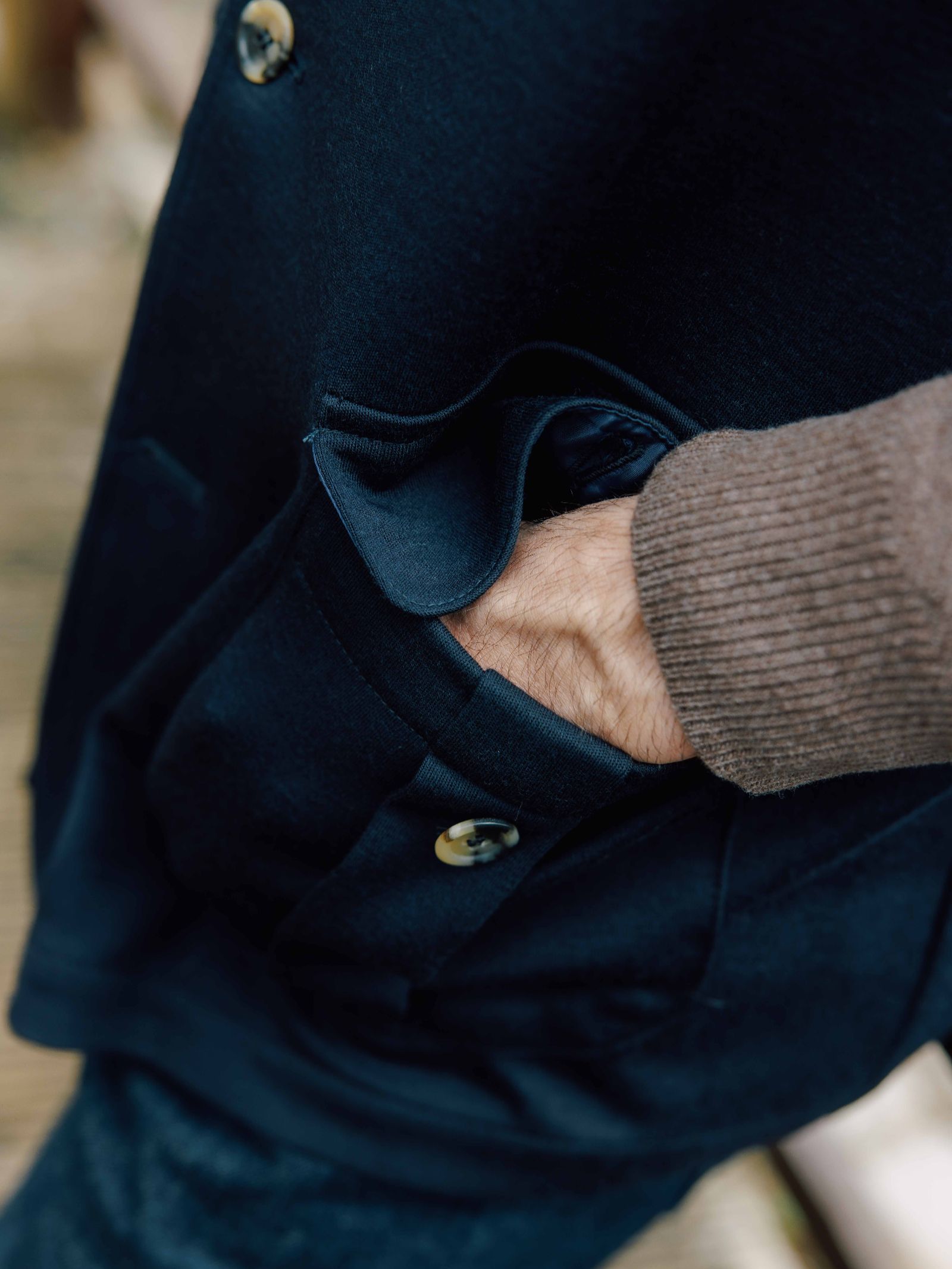 A close-up of a hand in the pocket of the Campbell's of Beauly Knitted Nehru Gilet, featuring two buttons and a pocket flap, with a brown ribbed sweater sleeve visible.