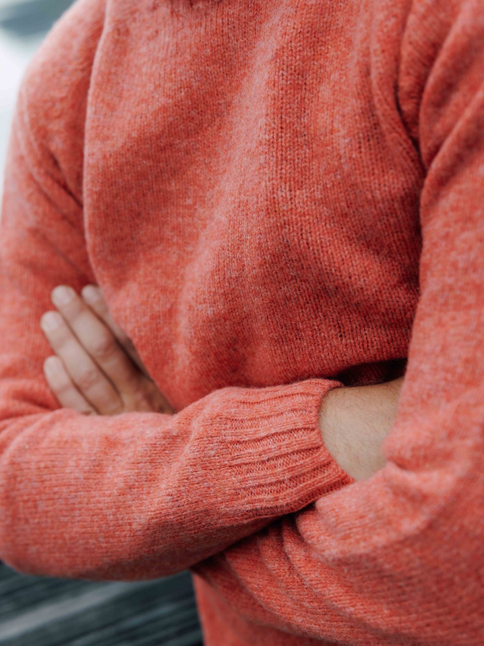 A person wearing the Campbell's of Beauly Shetland Jumper in orange-red lambswool stands with arms crossed. The photo highlights the sweater’s textured knit, focusing on the upper body without showing the face.