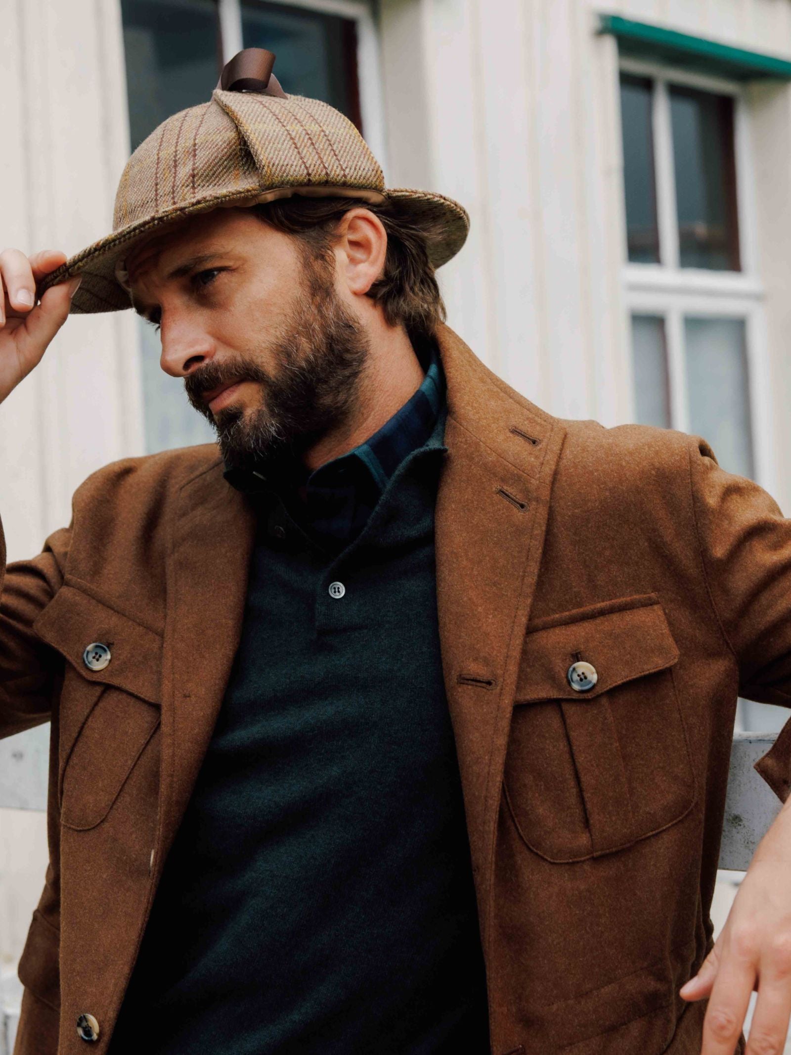 A bearded man adjusts a plaid hat while wearing a Campbell's of Beauly Field Teba Jacket over a dark shirt, standing in front of a building with white siding and large windows.