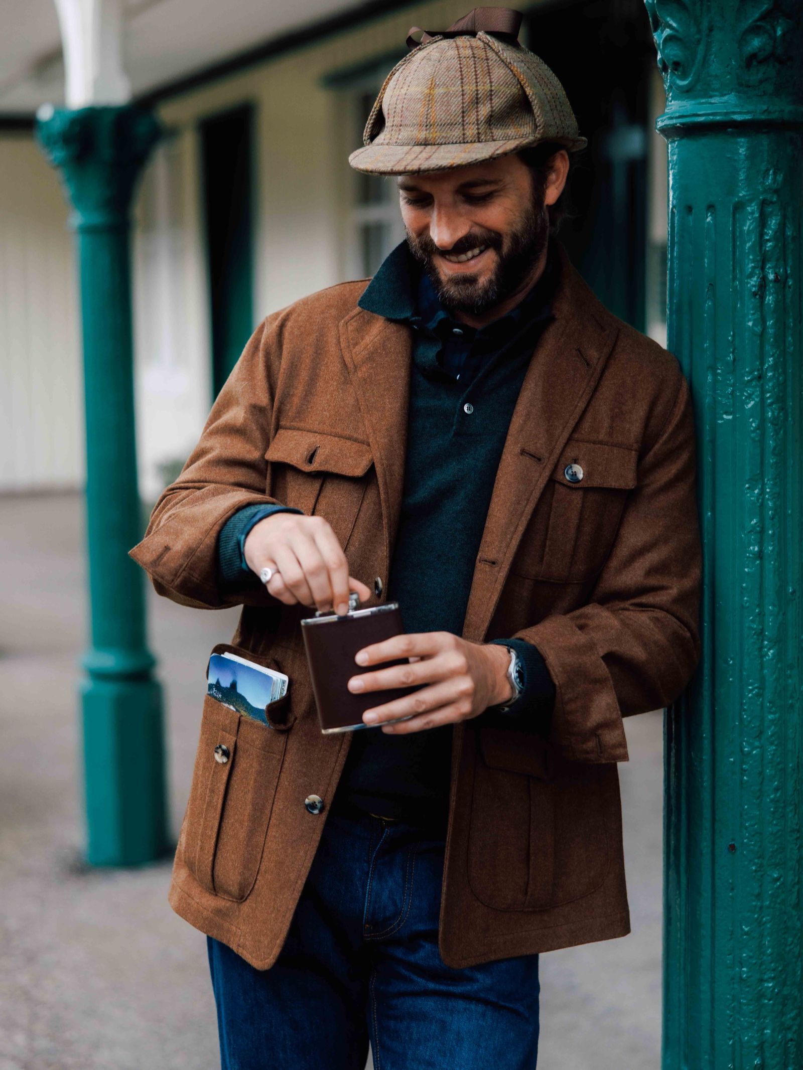 A smiling man in a Campbell's of Beauly Field Teba Jacket and plaid flat cap stands outdoors, leaning against a green pillar. He holds a metal flask, with a phone visible in his jacket pocket.