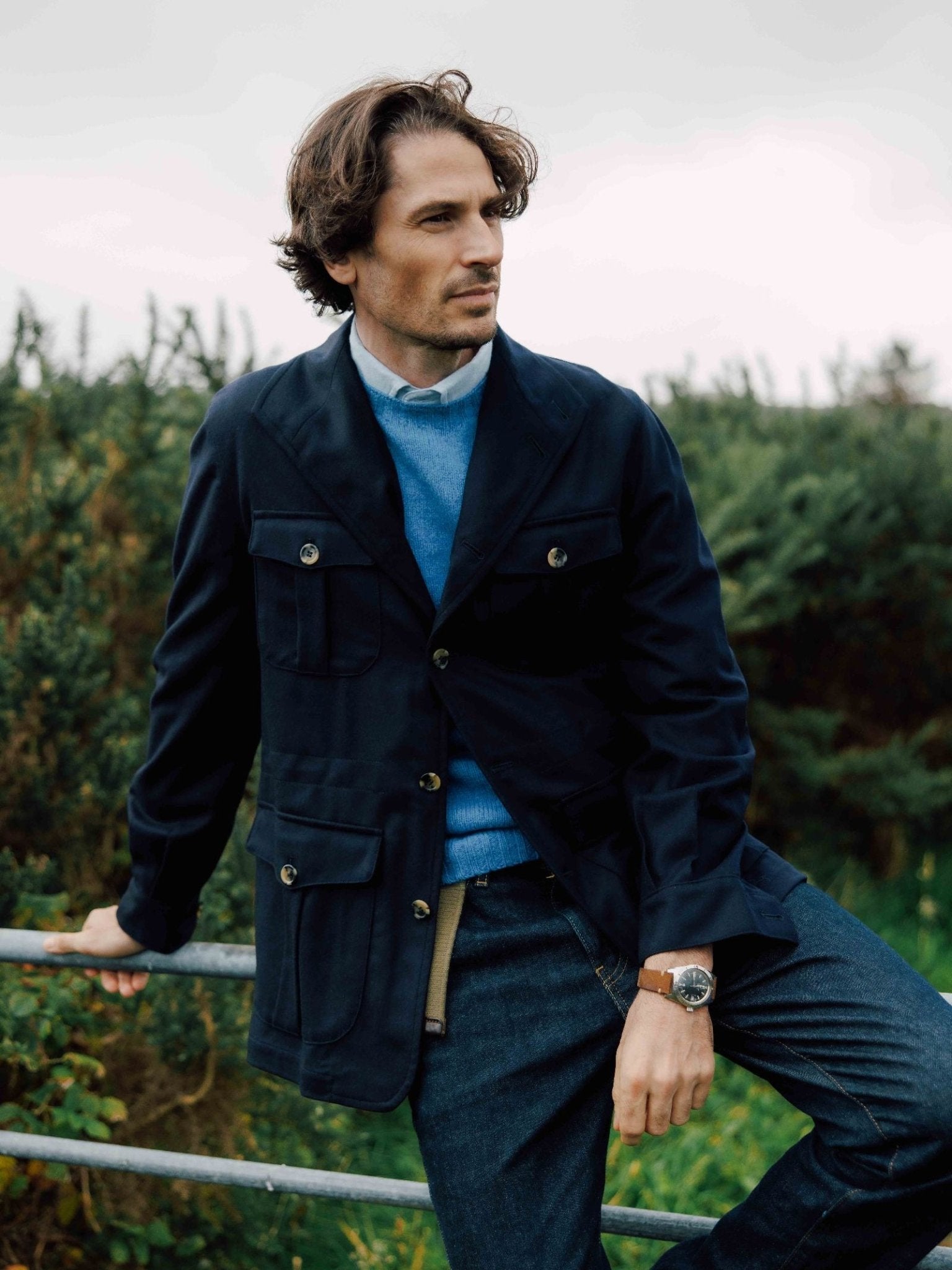 A man with wavy brown hair wears the Campbell's of Beauly Auld Stock Field Teba Jacket, a blue sweater, and jeans as he leans casually on a metal fence outdoors, surrounded by greenery and bushes.
