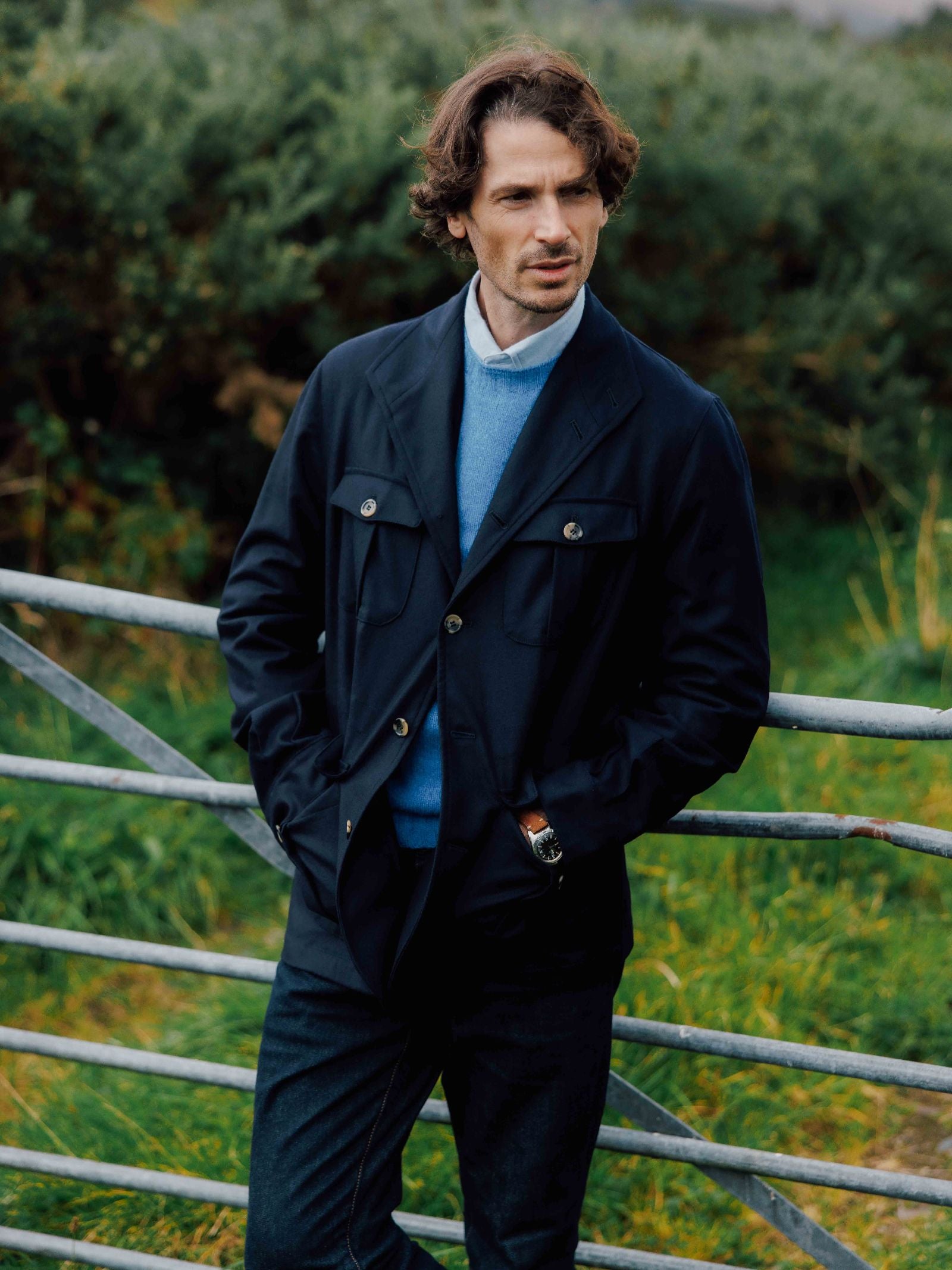 A man with wavy brown hair stands outdoors by a metal fence, hands in pockets, wearing Campbell's of Beauly Field Teba Jacket over a blue sweater and white shirt. Greenery fills the background.