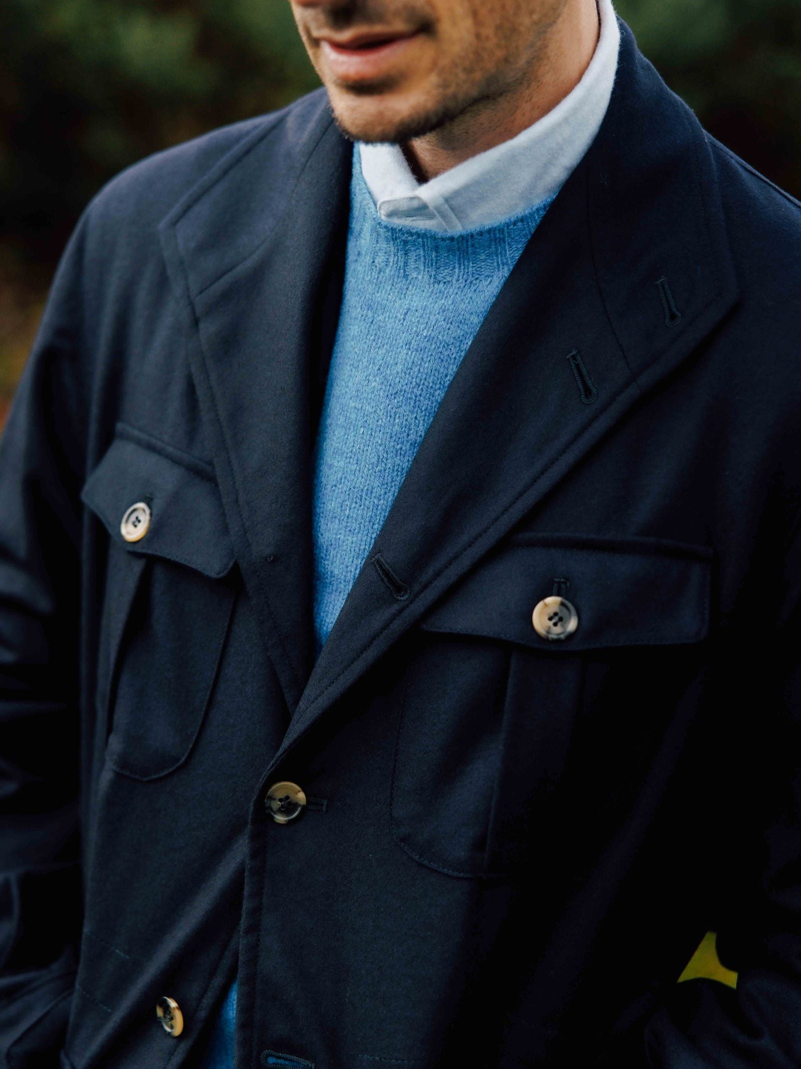 A man wears the Campbell's of Beauly Field Teba Jacket in navy blue with large buttons, layered over a light blue sweater and white collared shirt, shown from shoulders to mouth, standing outdoors.