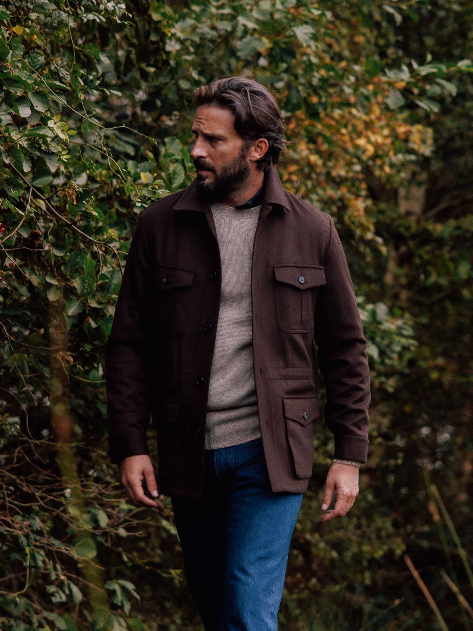 A man with dark hair and a beard strolls outdoors near green foliage, wearing a Campbell's of Beauly Winter Aigas Field Jacket over a beige sweater and blue jeans.
