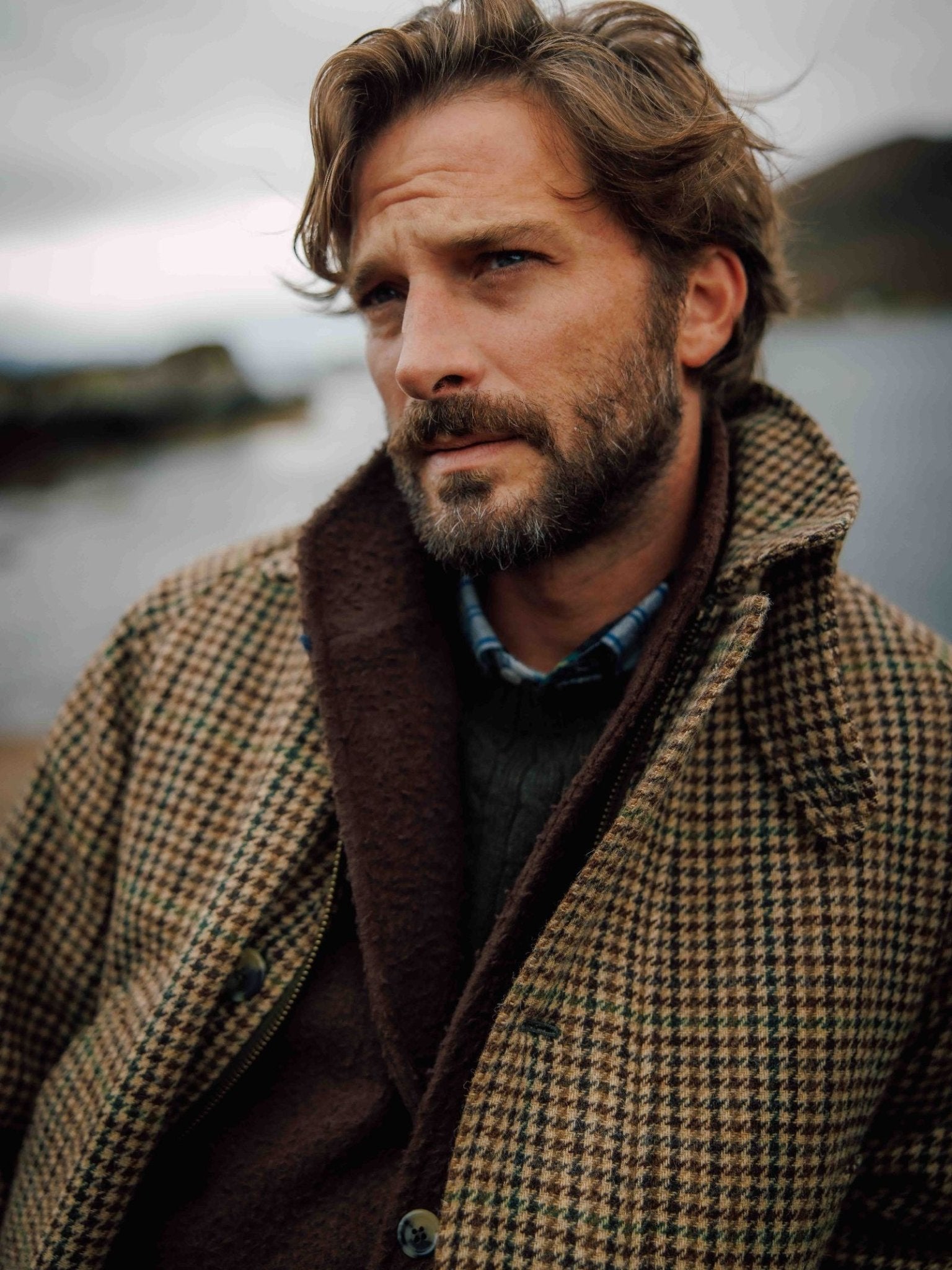 A man with wavy brown hair and a beard gazes into the distance, standing outdoors by water on a cloudy day, wearing the Campbell's of Beauly Cannich Coat over a dark sweater.