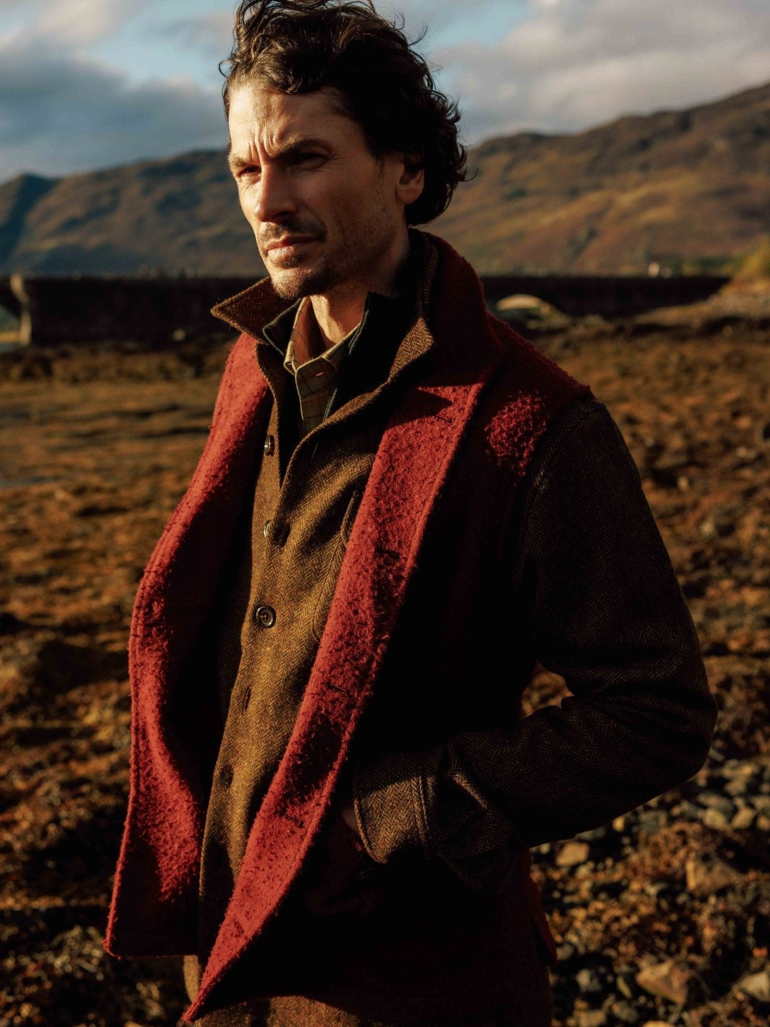 A man stands outdoors in rugged terrain wearing the Campbell's of Beauly Casentino Fleece Gilet, featuring a brown body with a dark red collar and lapels. He gazes off to the side, mountains and a cloudy sky in the background.