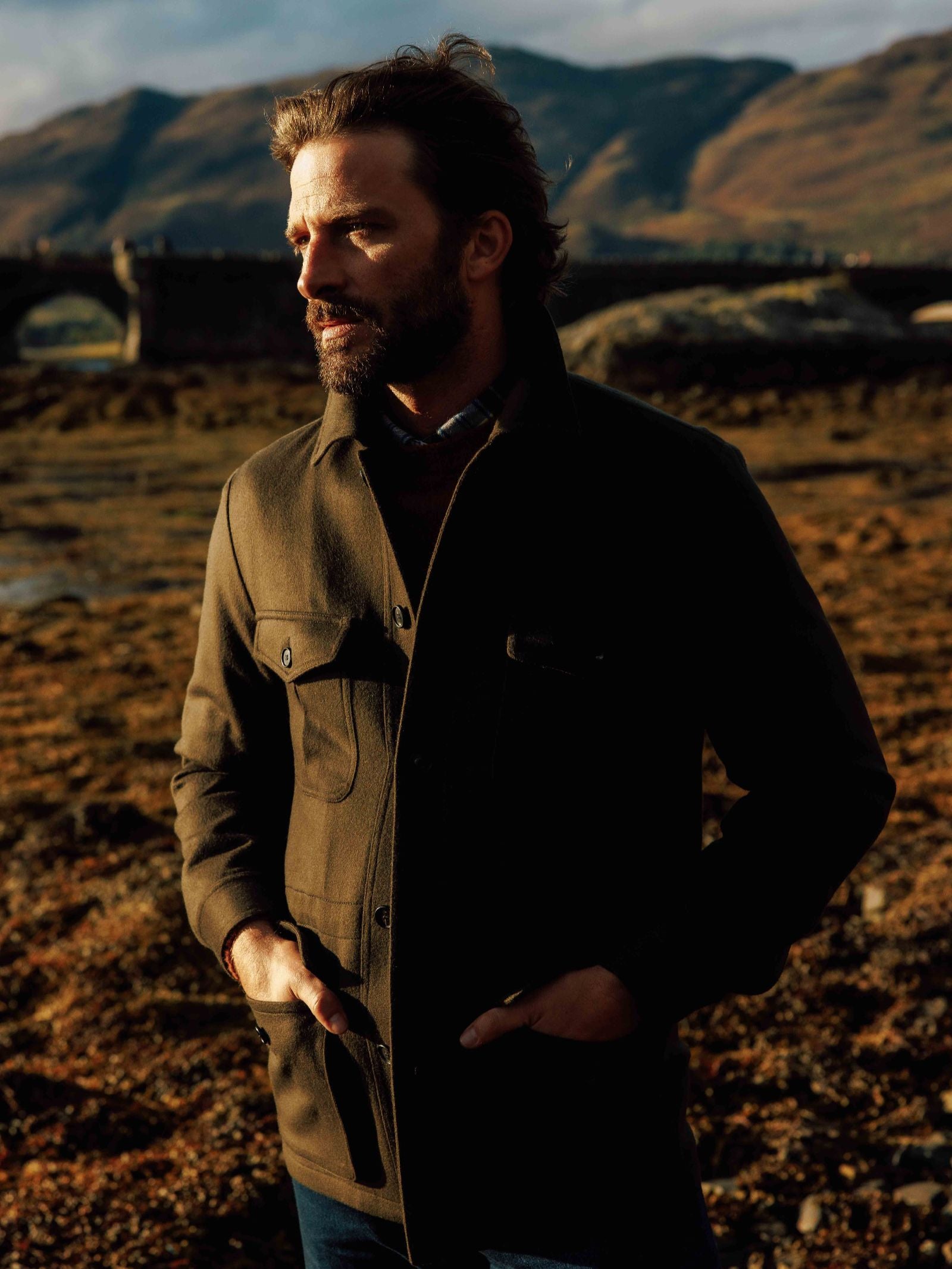 A bearded man stands outdoors in warm light, wearing a green Campbell's of Beauly Winter Aigas Field Jacket with his hands in his pockets. Behind him are mountains and a stone bridge under a partly cloudy sky.
