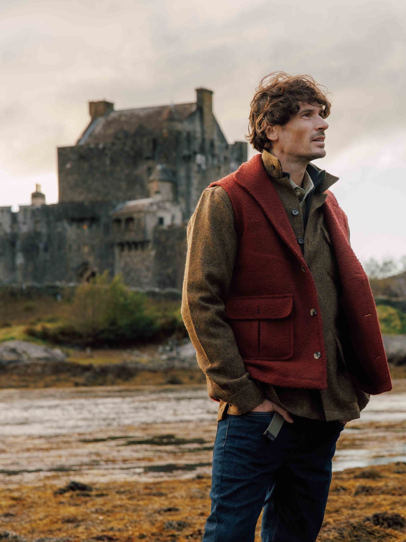 A man stands outdoors on rocky ground with an old stone castle behind him, wearing a red Casentino Fleece Gilet by Campbell's of Beauly over a brown jacket beneath a cloudy sky.