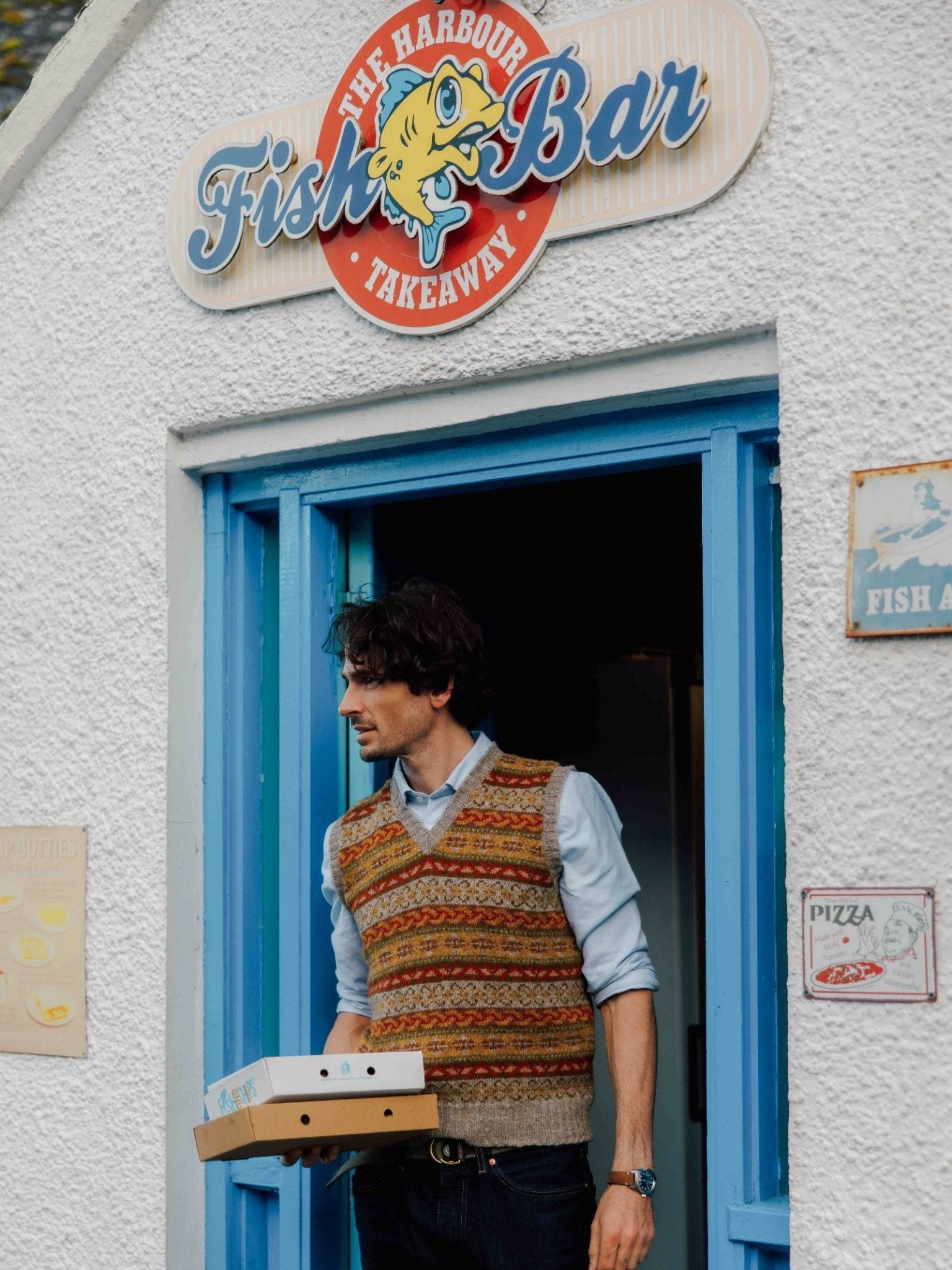 A man in a Campbell's of Beauly Fairisle Slipover stands at the doorway of a white building with blue trim, holding two pizza boxes beneath The Harbour Fish Bar Takeaway sign.