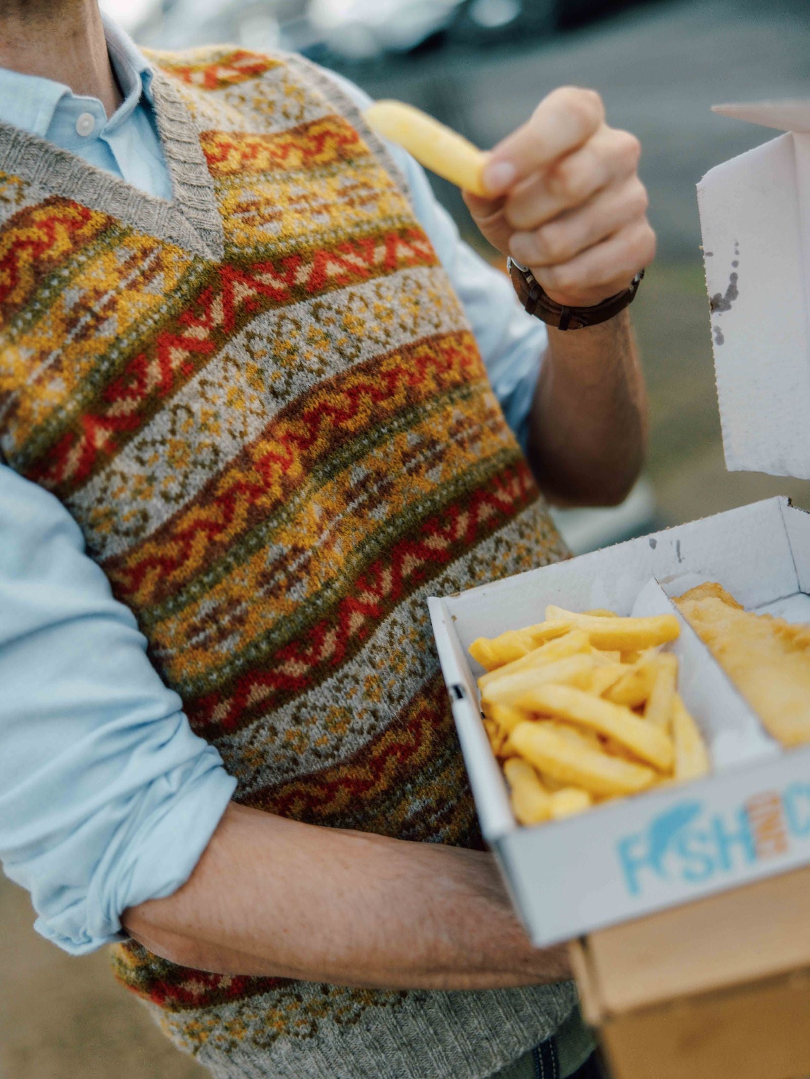 Wearing a Campbell's of Beauly Fairisle Slipover, a person outdoors holds a "FISH and CH" box of fish and chips, raising a fry to their mouth. The background is slightly blurred.