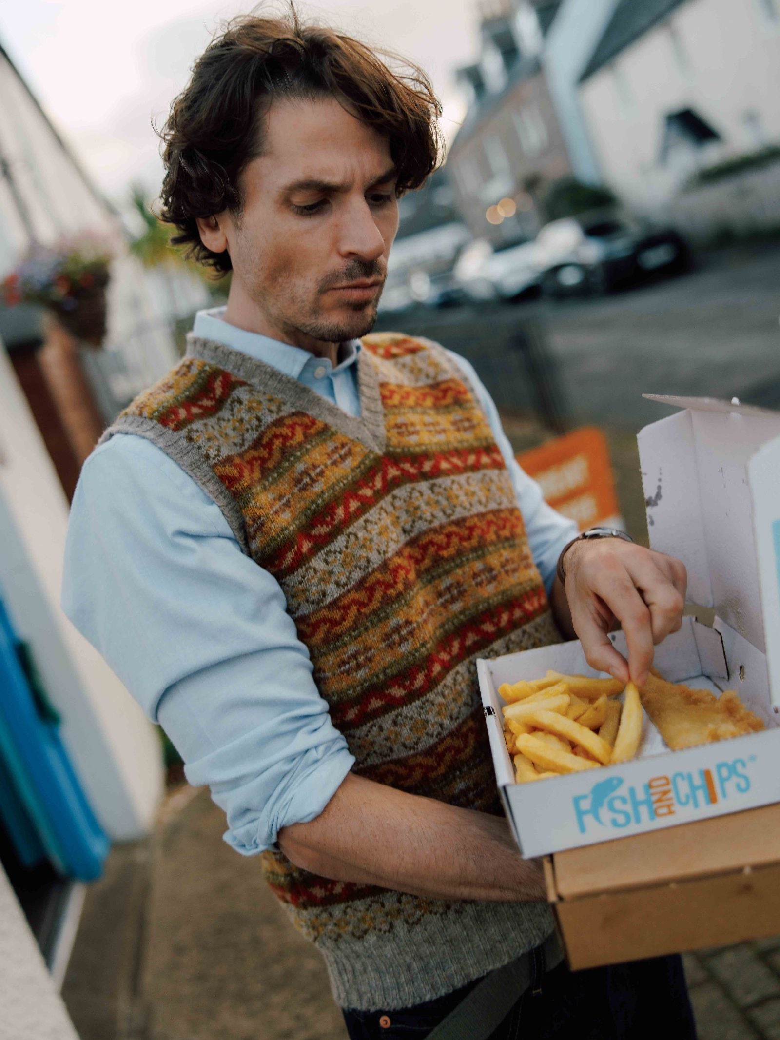 A man wearing the Campbell's of Beauly Fairisle Slipover stands on a street with houses and cars in the background, holding a box of fish and chips and picking up a fry.