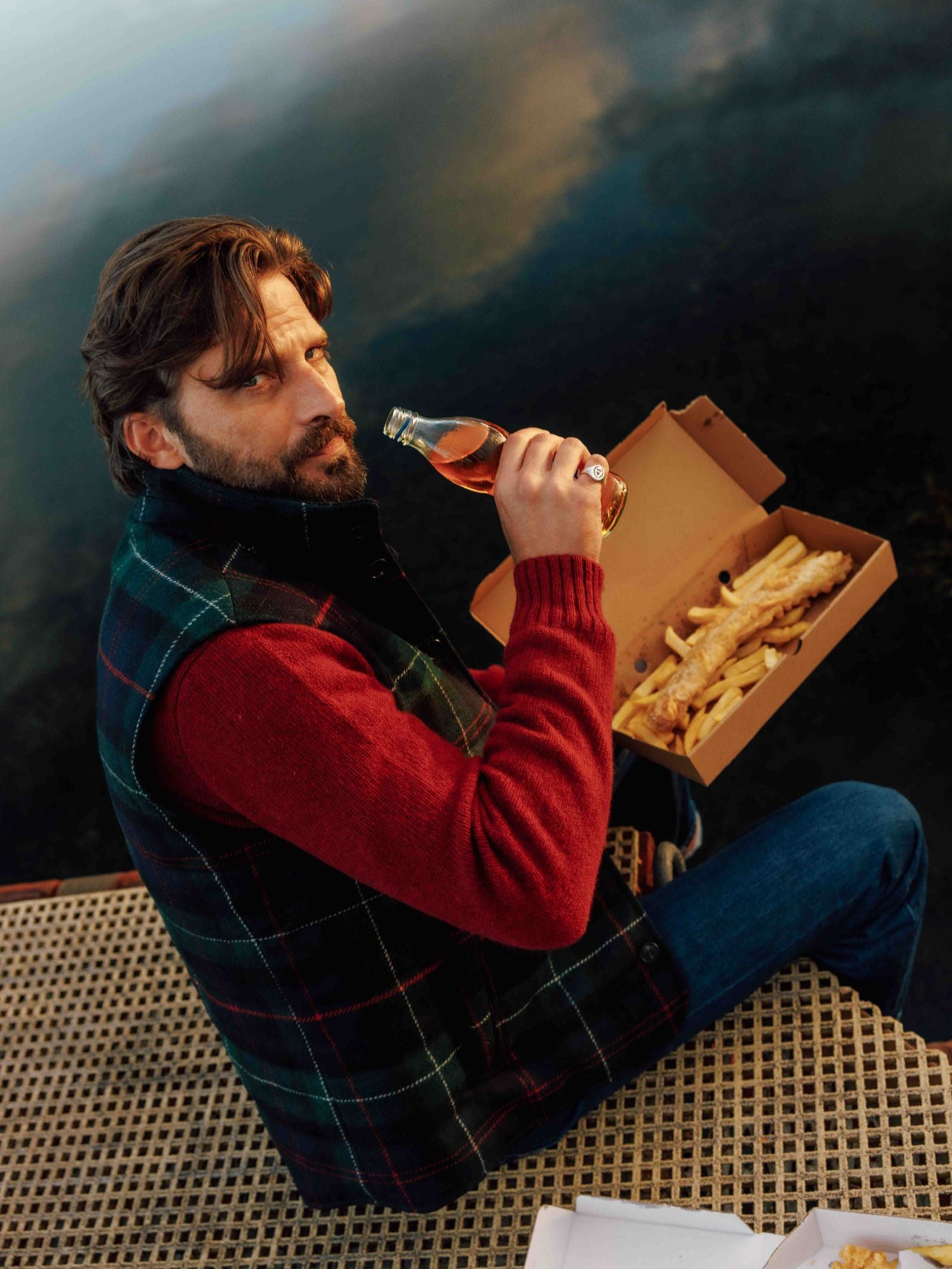 A man with brown hair and a beard, wearing a red sweater and the Campbell’s of Beauly Auld Stock Quilted Tweed Buttoned Gilet, sits on a dock by the water holding fries and a bottle, looking up at the camera.