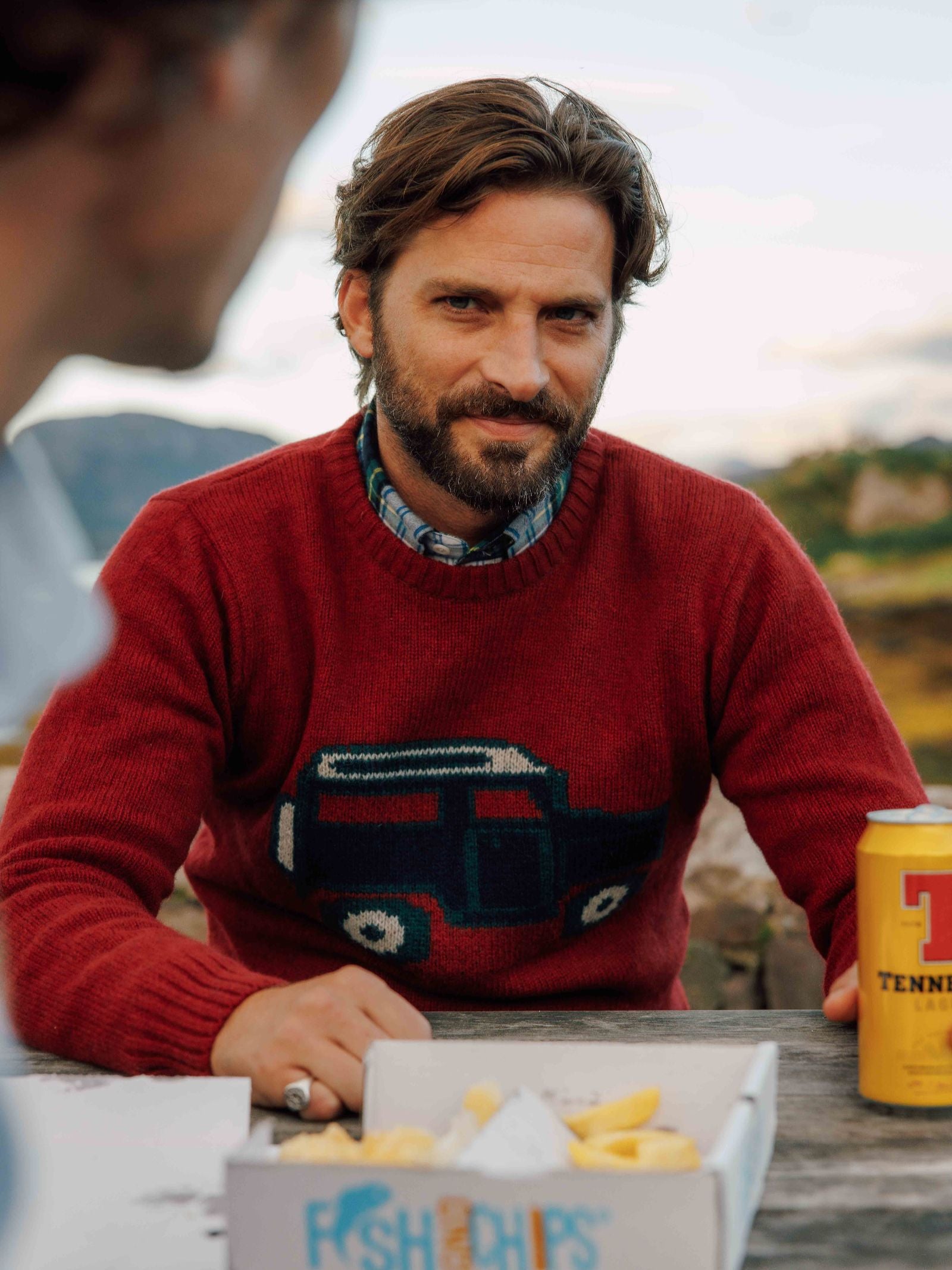 A bearded man wearing the Campbell's of Beauly Land Rover Defender Jumper sits outdoors at a picnic table with a food box of fish and chips and a yellow can of Tennent's beer in front of him, gazing off-camera.