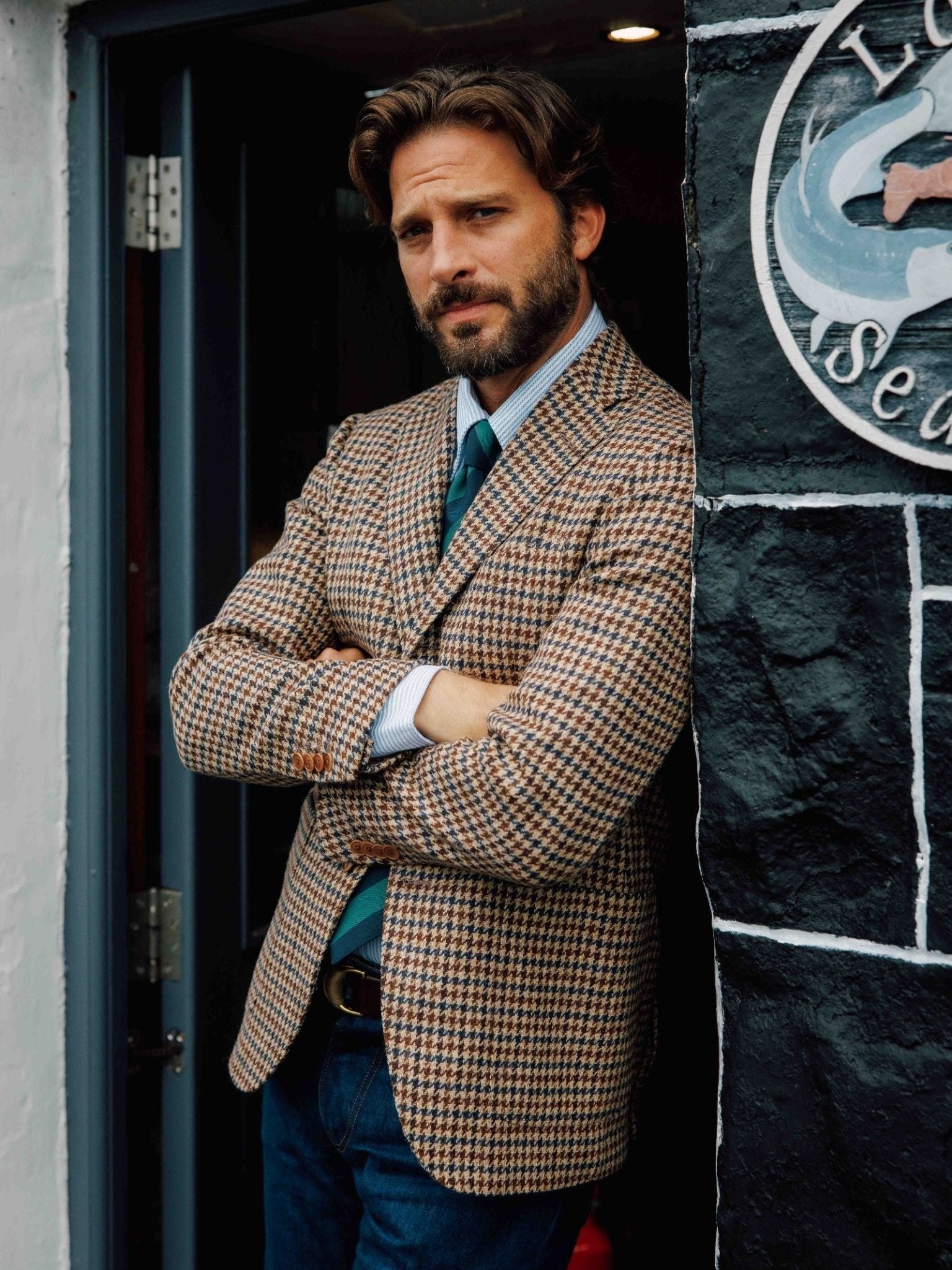 A man with wavy brown hair and a beard stands in a doorway, arms crossed, wearing the Campbell's of Beauly Navy/Brown Gun Club Tweed Jacket over a shirt and tie with blue jeans. A partial circular sign appears on the black wall next to him.