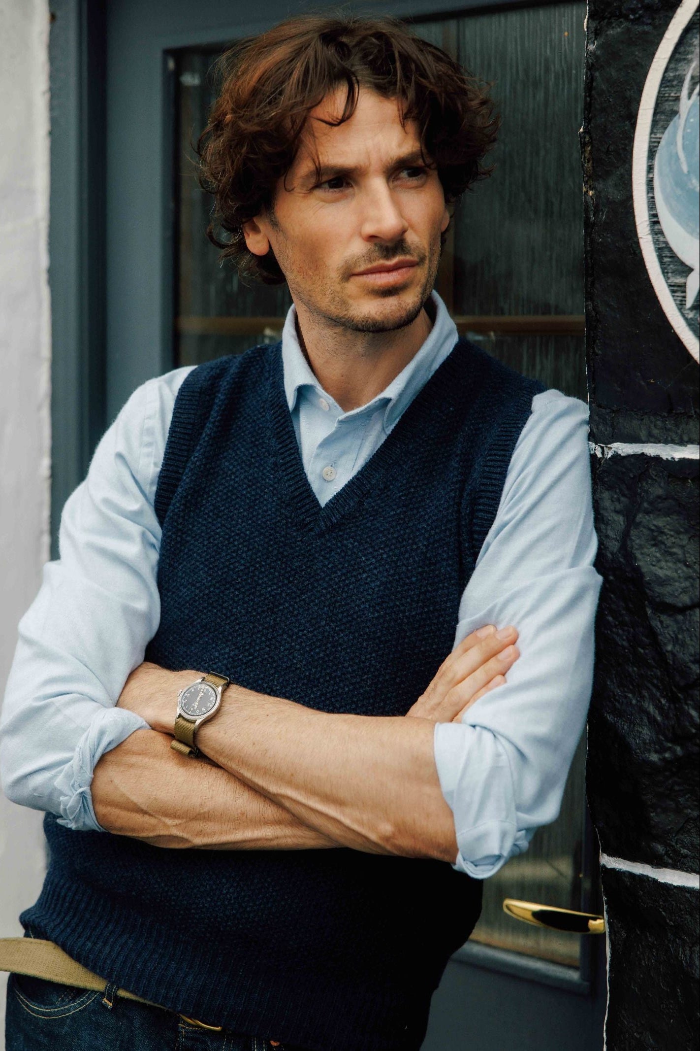 A man with wavy brown hair, wearing a Campbell's of Beauly Moss Stitch Knitted Slipover made in the UK over a light blue shirt, stands with arms crossed, leaning against a door. He wears a watch and gazes thoughtfully to the side.